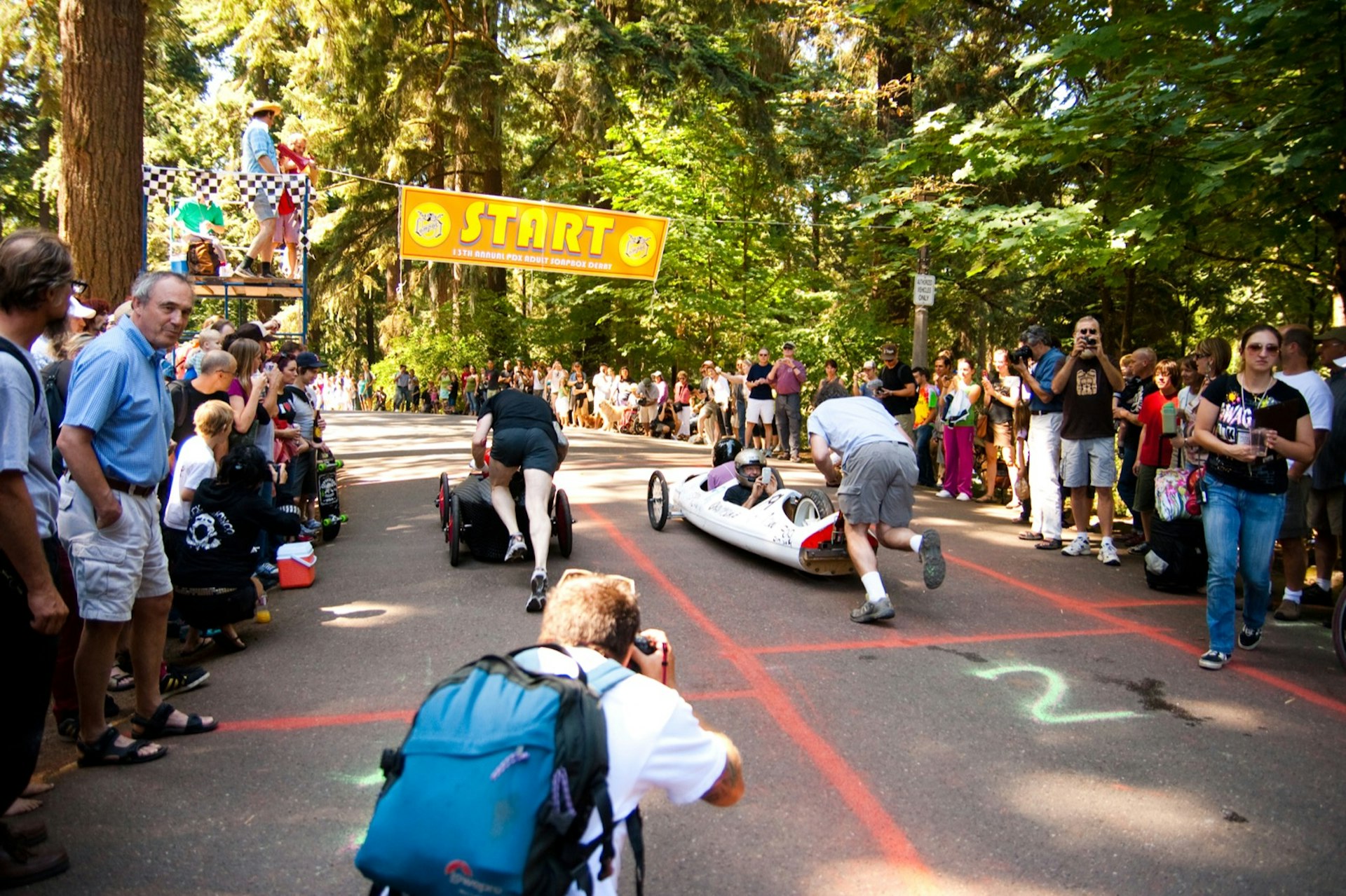 People push very sleek soapbox cars through a forested area on a road with lanes painted on it