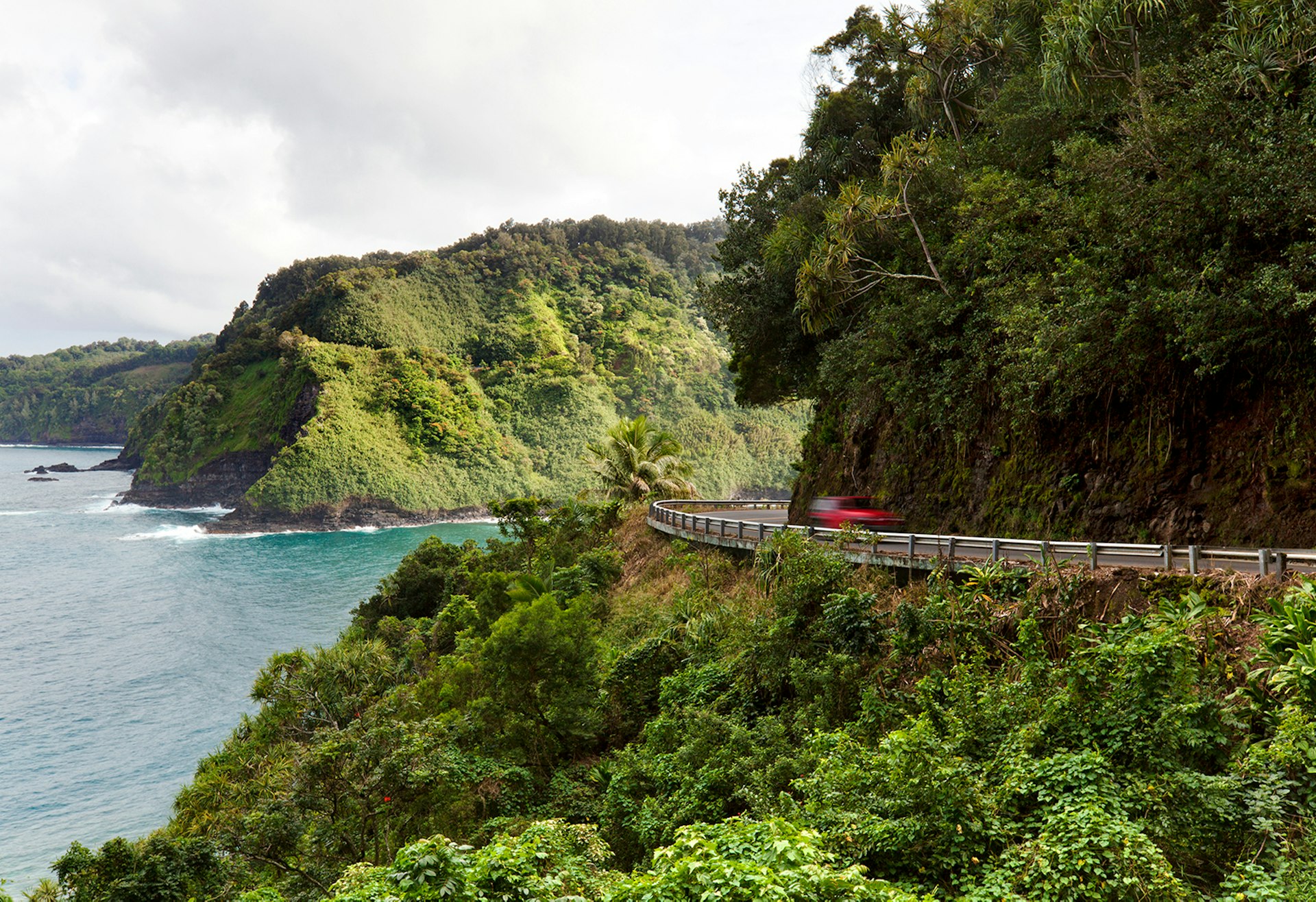 A car drives along a coastal road lined in tropical vegetation; maui