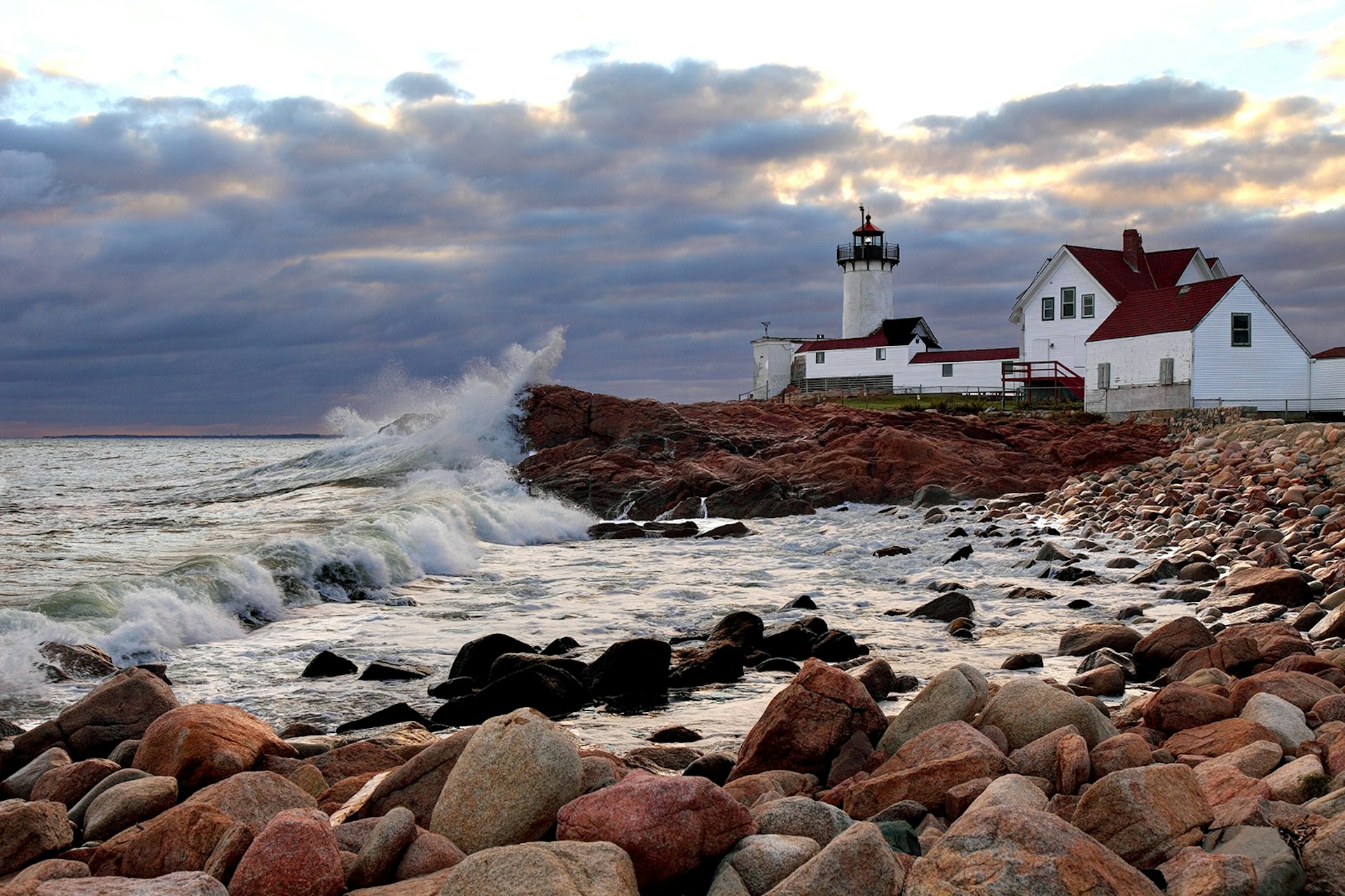 Waves crash beneath a lighthouse, with a rocky shore in the foreground