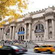 Cars and taxis pass by the facade of a museum as people congregate on the steps