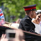 Above a blur of waving hands from gathered crowds is the open-topped carriage carrying Prince Harry and Meghan Markle; Harry in uniform looks forward while Meghan has her head turned towards to crowd and camera, with a bright smile and wave; a blurred British flag flies in the background against a forested backdrop © DANIEL LEAL-OLIVAS / Contributor / Getty Images