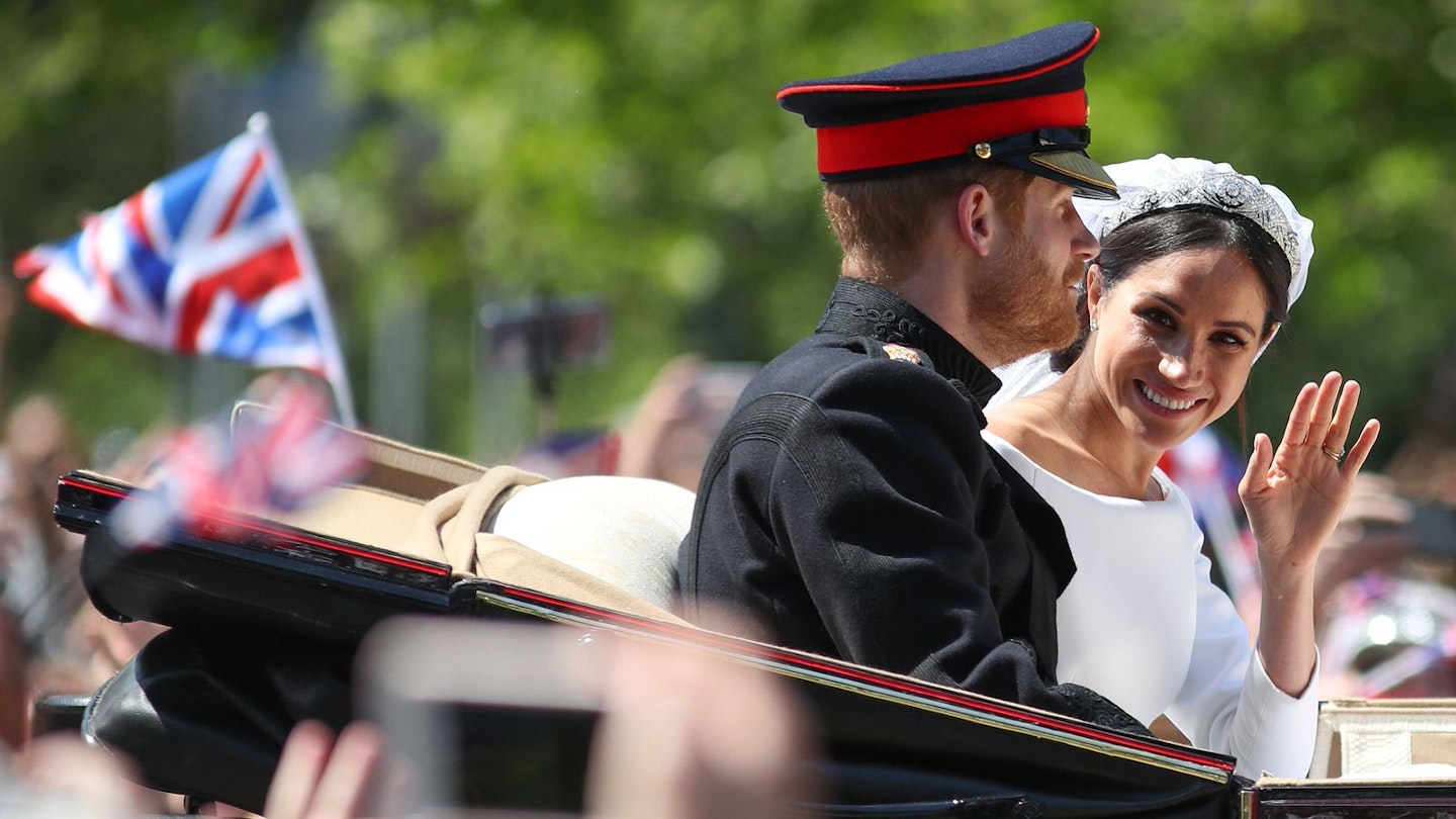 Above a blur of waving hands from gathered crowds is the open-topped carriage carrying Prince Harry and Meghan Markle; Harry in uniform looks forward while Meghan has her head turned towards to crowd and camera, with a bright smile and wave; a blurred British flag flies in the background against a forested backdrop © DANIEL LEAL-OLIVAS / Contributor / Getty Images