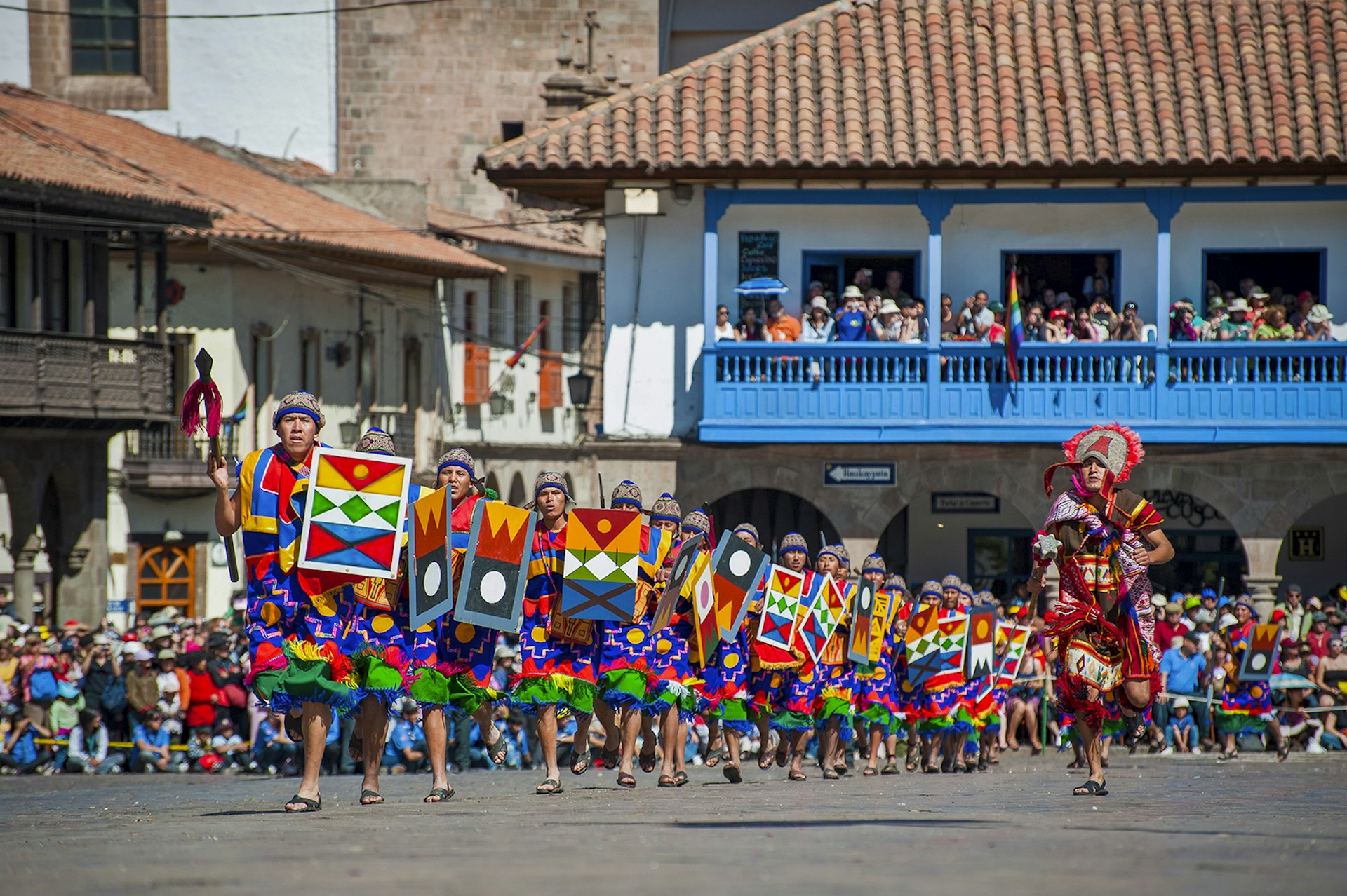 A line of men wearing colorful traditional Inca dress holding shields with geometric patterns walk through historic Cuzco, Peru for the solstice festival Inti Raymi.