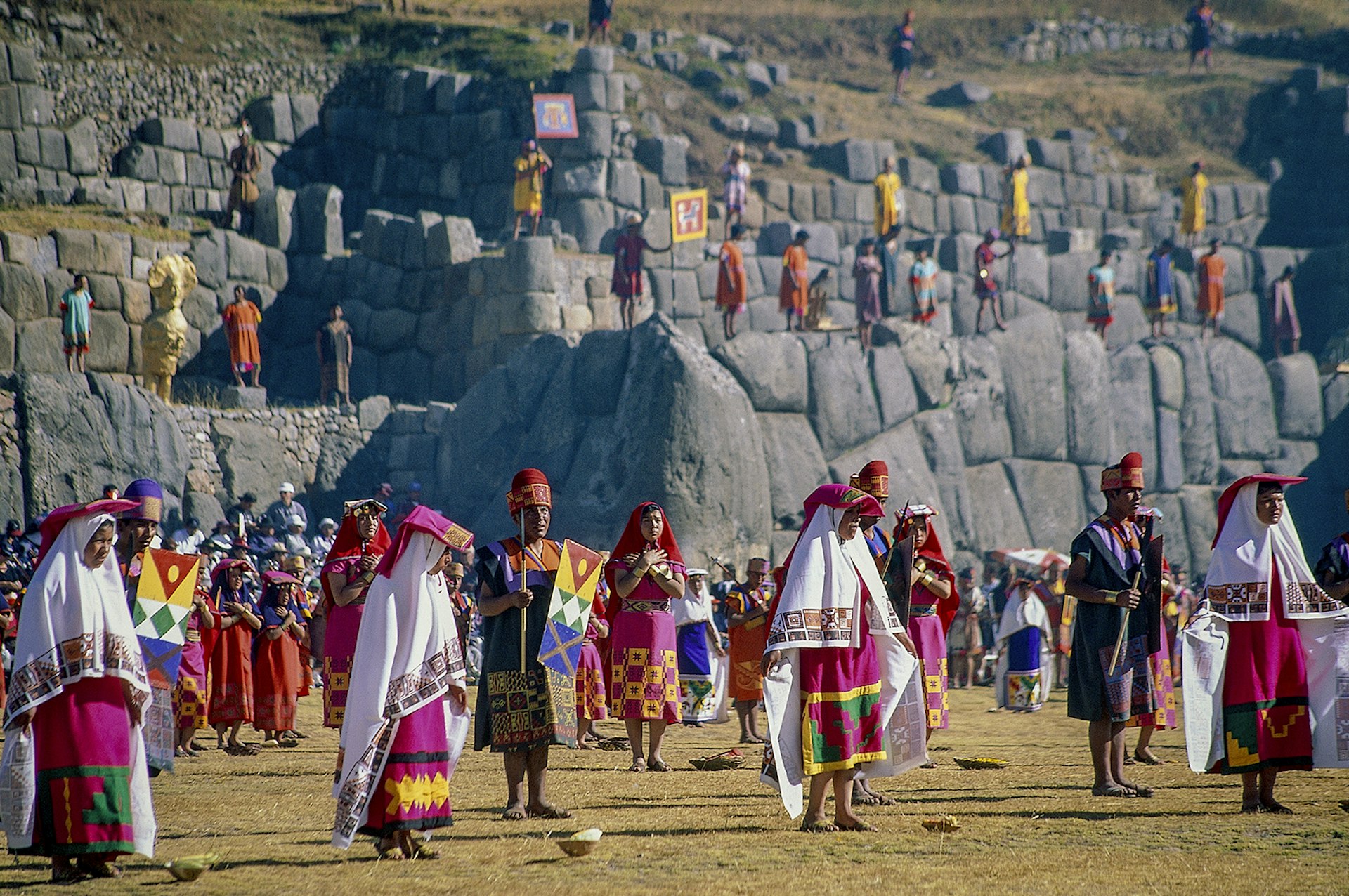 Men and women in colorful Inca dress stand in the main plaza of Sacsayhuaman, with its large stone walls visible in the background. Cuzco, Peru.