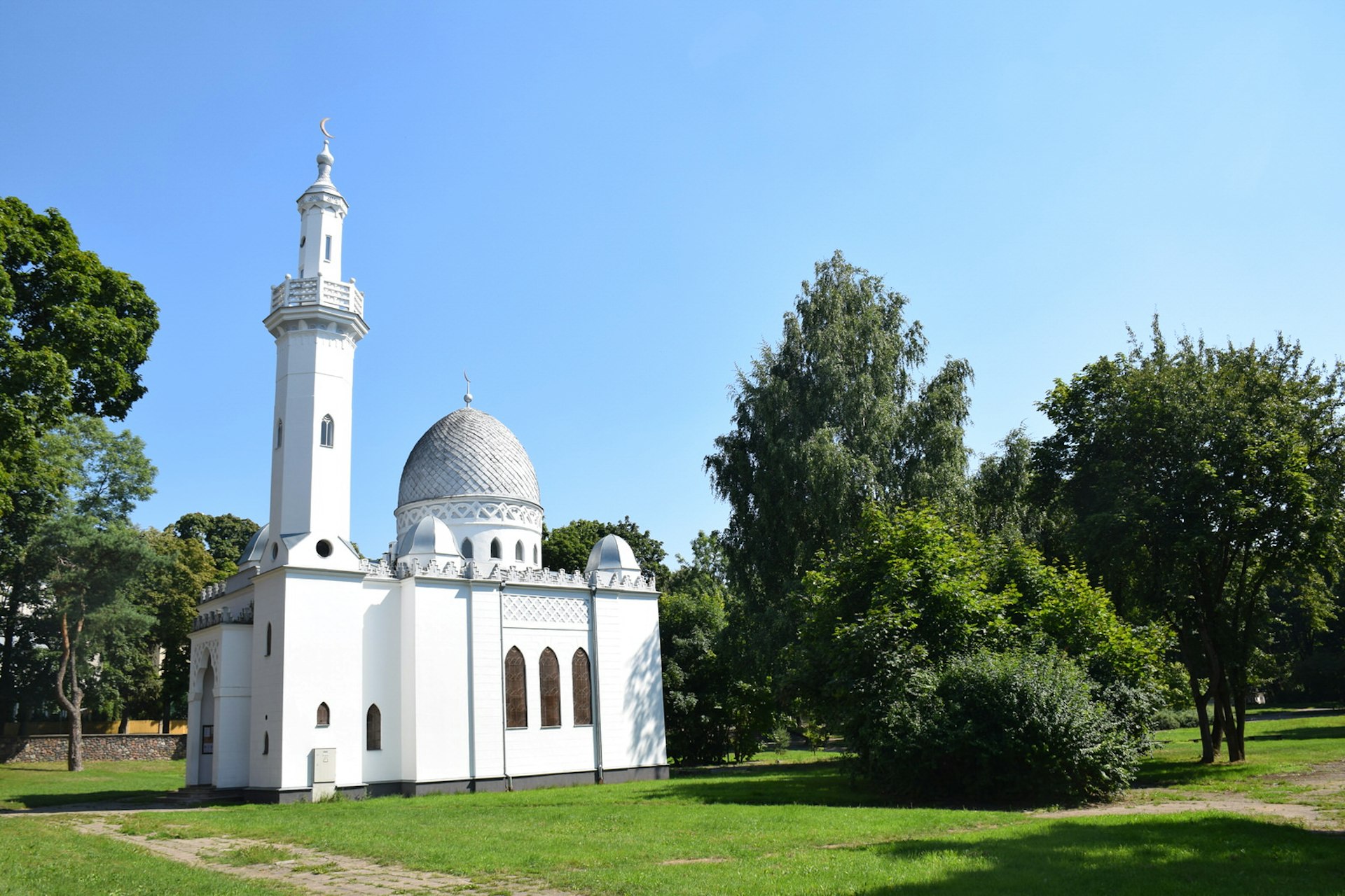 A bright, white mosque with dome and elegant minaret sitting amidst dark green trees