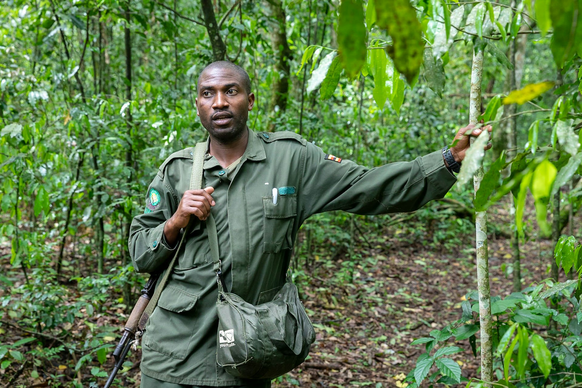 Africano, the Ugandan park guide, stands dressed in his green uniform, with one arm resting on a tree, the other holding the shoulder strap of his rifle © Bella Falk
