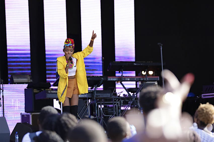 Nairobi June - A female musician stands on stage, with her right hand holding the microphone to her face, the left raised high with her index finger pointing skyward; she's wearing a bright yellow jacked and red hat; the blurred heads of the audience are in the foreground
