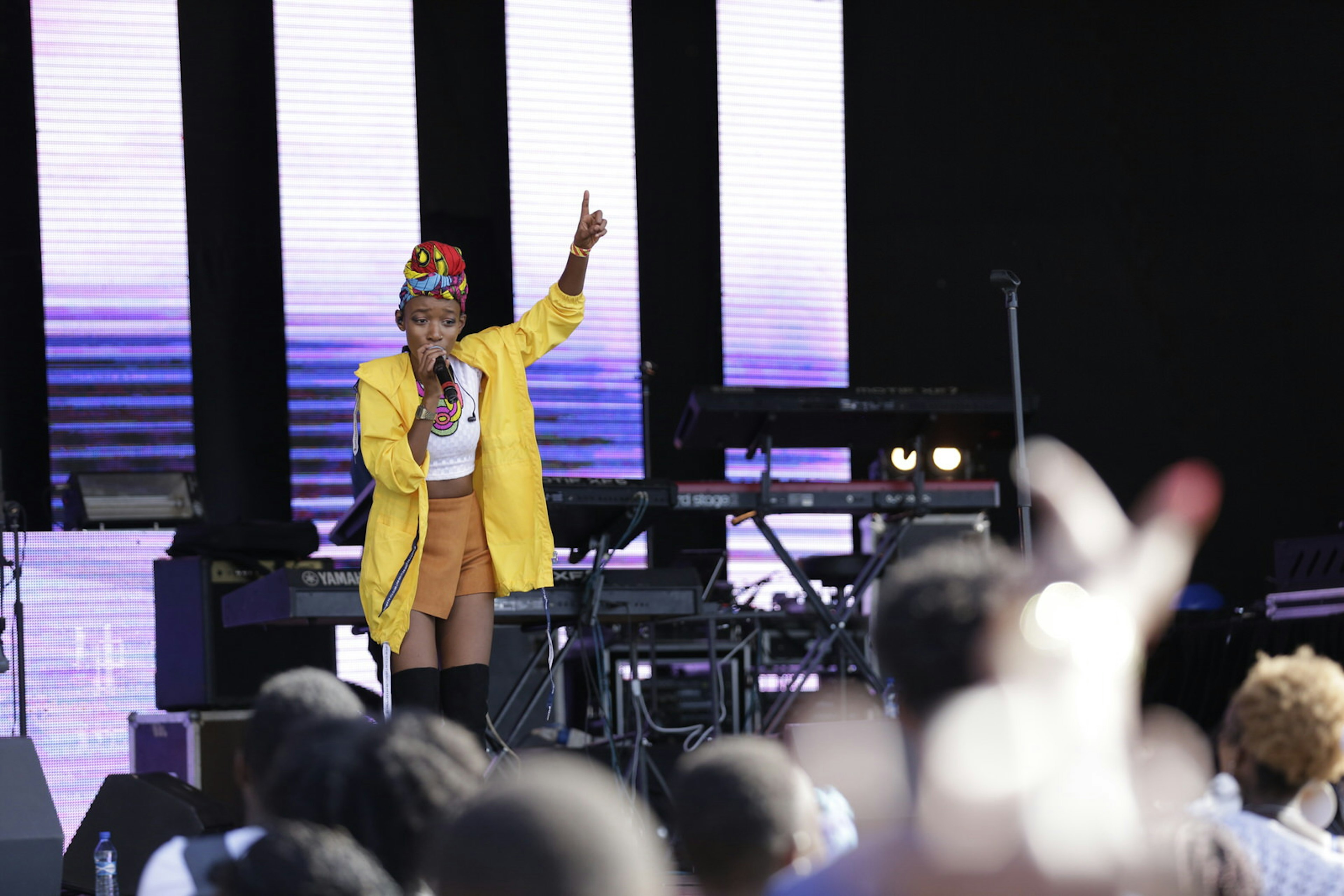A female musician stands on stage, with her right hand holding the microphone to her face, the left raised high with her index finger pointing skyward; she's wearing a bright yellow jacked and red hat; the blurred heads of the audience are in the foreground