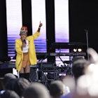 A female musician stands on stage, with her right hand holding the microphone to her face, the left raised high with her index finger pointing skyward; she's wearing a bright yellow jacked and red hat; the blurred heads of the audience are in the foreground