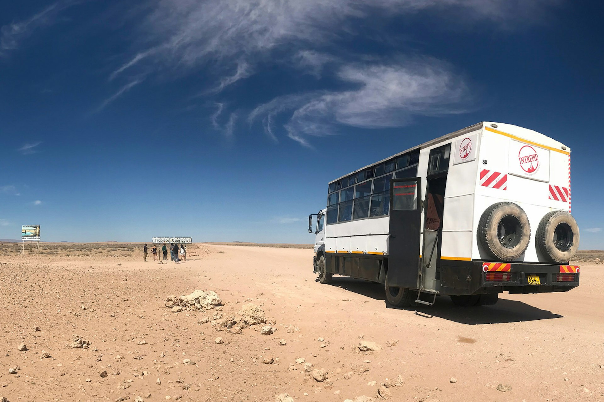 The large overland truck sits in the foreground with its door open, with half a dozen people standing in the distance next to the Tropic of Capricorn roadsign; the setting is a dusty, rocky desert with a deep blue sky and whispy clouds @ Sarah Reid / Lonely Planet