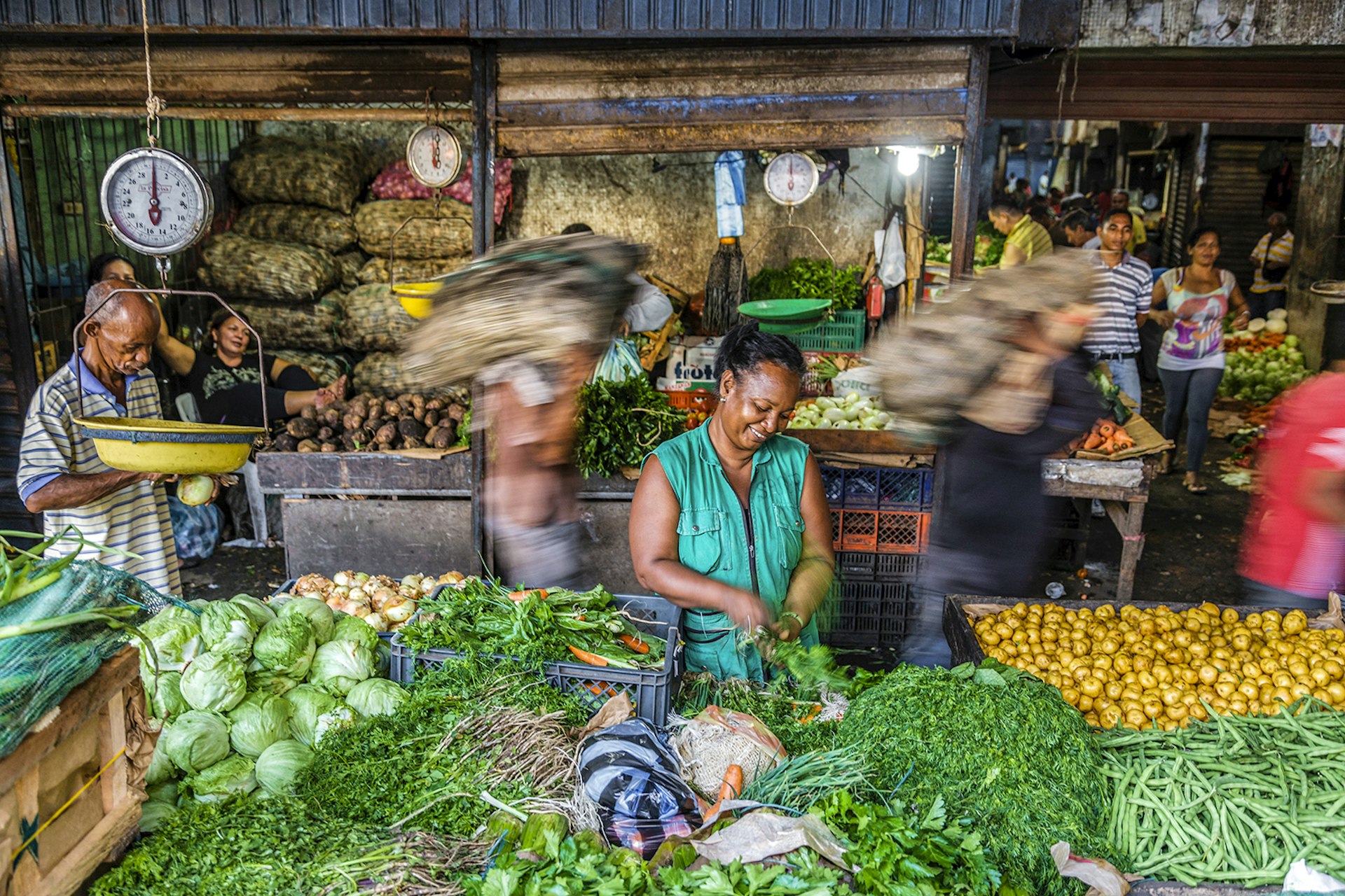 A smiling woman tends to green vegetables at a stand in a Colombian market. Men pass behind her carrying bags of produce on their shoulders.