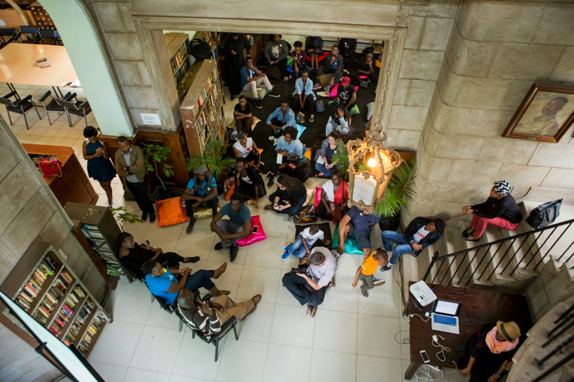 Nairobi June - Shot looking down from a high balcony, a group sits tightly packed on a library floor in front of three speakers sitting in chairs