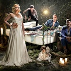 Celebrities West End Braodway - Gwendoline Christie leads the cast of A Midsummer Night's Dream at the Bridge Theatre, London. Gwendoline stands in front of the other cast members, posed around a wrought iron bed, in a floor-length, white silk gown © Perou