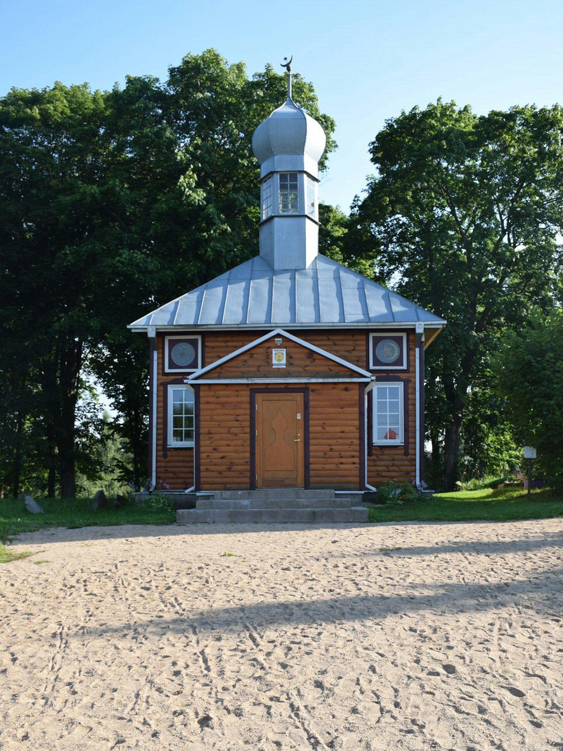 A small simple mosque made from light wood, with a central minaret topped with an onion dome and crescent