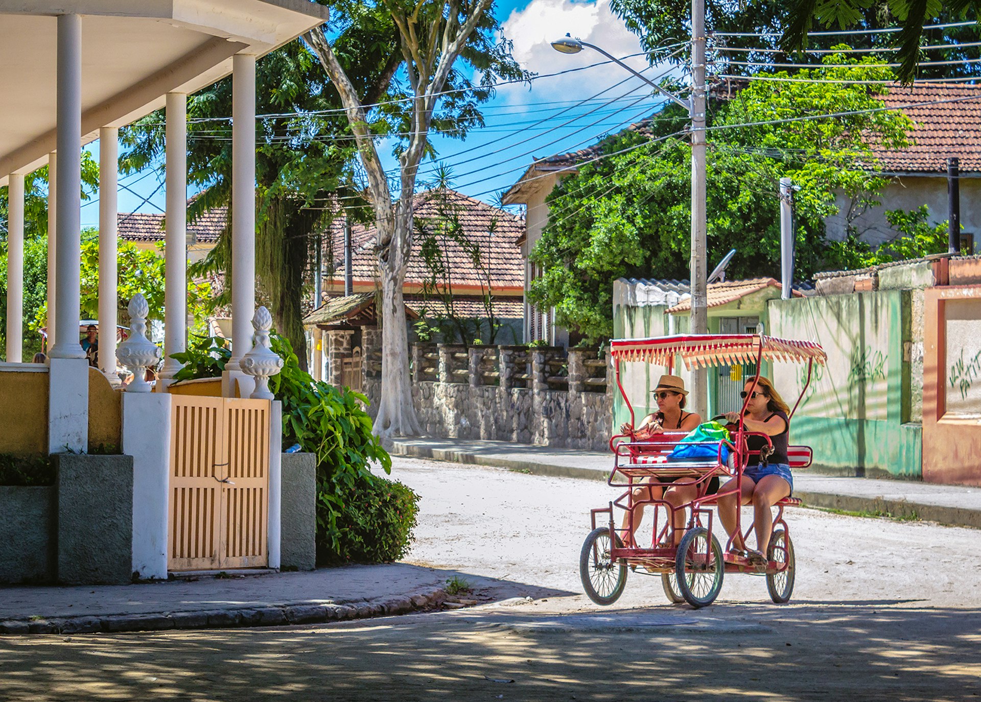 Two women pedal a foot-powered cart through a street lined with pastel-colored buildings on Ilha da Paquetá, Brazil