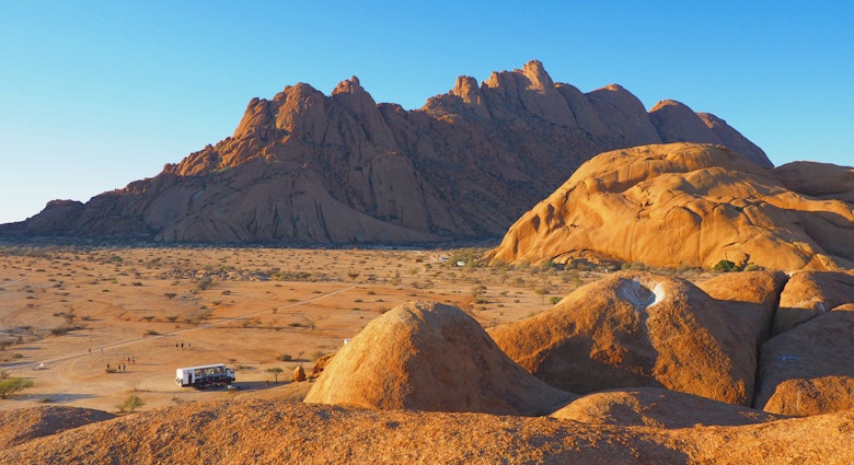 A large overland truck looks much like a piece of lego as it sits on the red Namibian soil surrounded by massive, bulbous rock monoliths at Spitzkoppe @ Sarah Reid / Lonely Planet