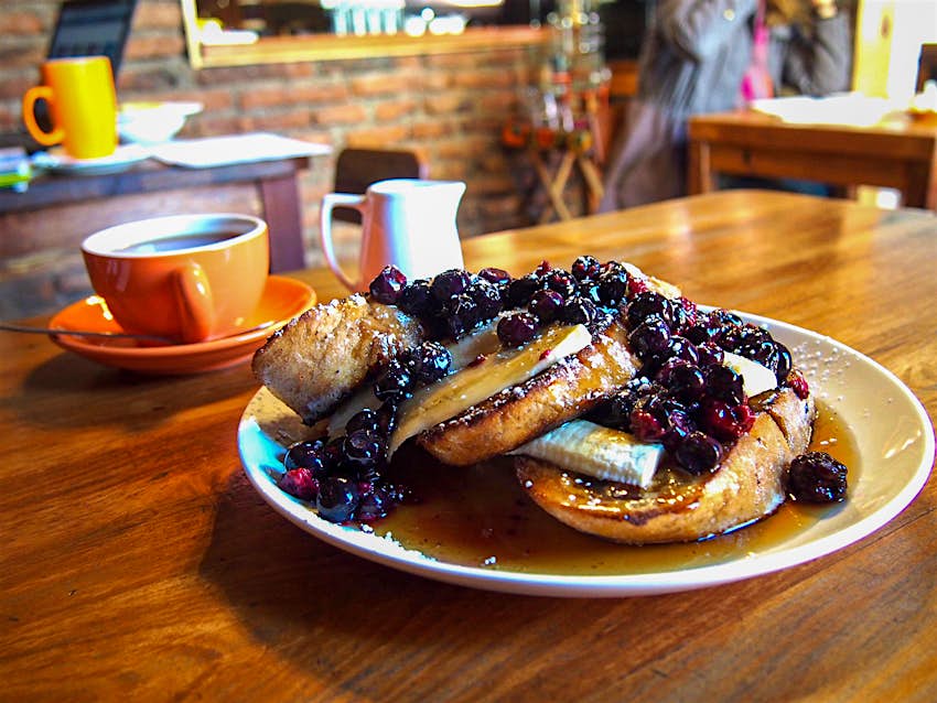 A plate with french toast topped with bananas and blueberries, with a cup of coffee in the background. Santiago, Chile.
