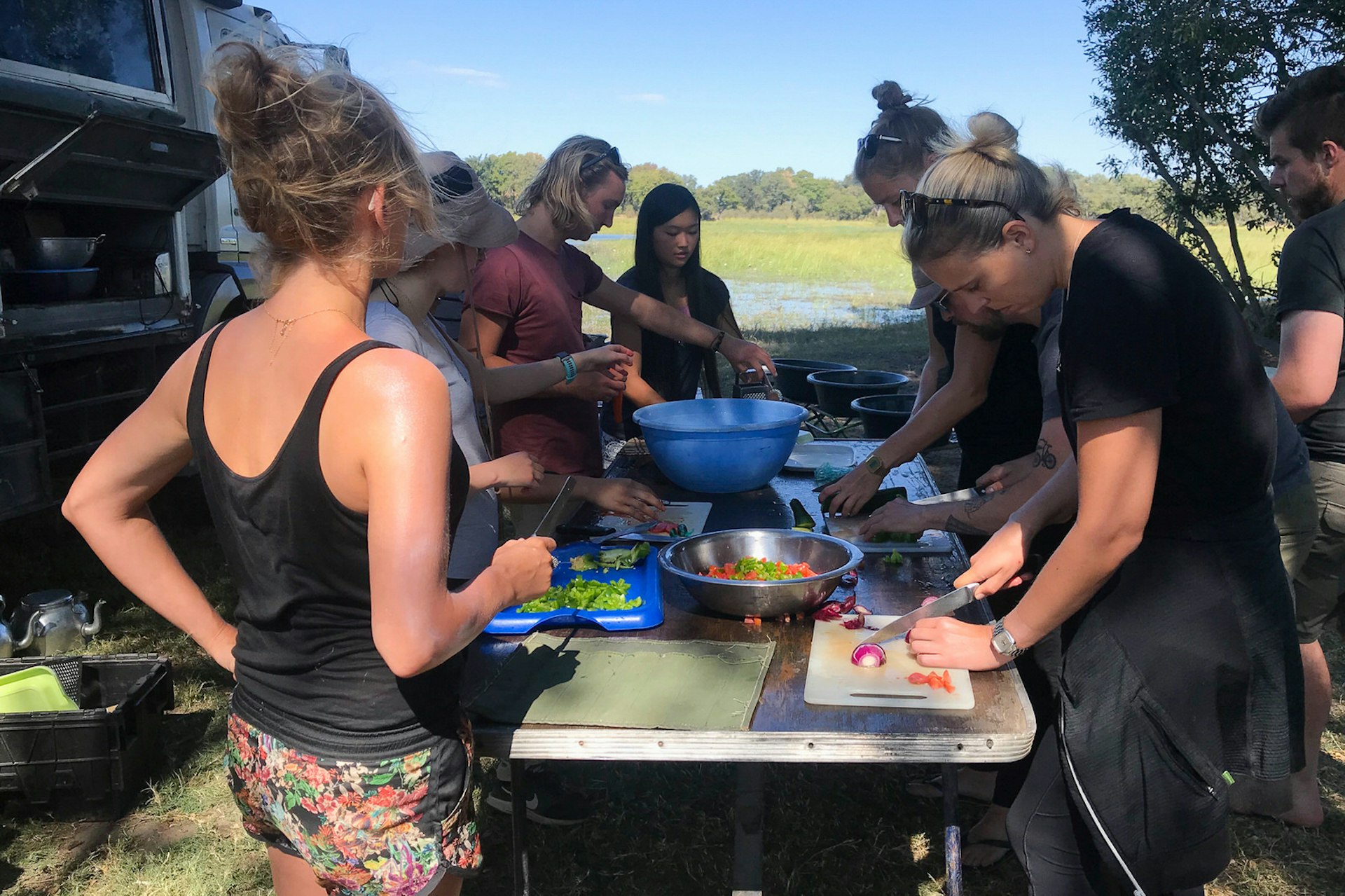Half a dozen people are standing around a portable table, each preparing something different for an upcoming meal; the overland truck sits next to them in the wilderness @ Sarah Reid / Lonely Planet
