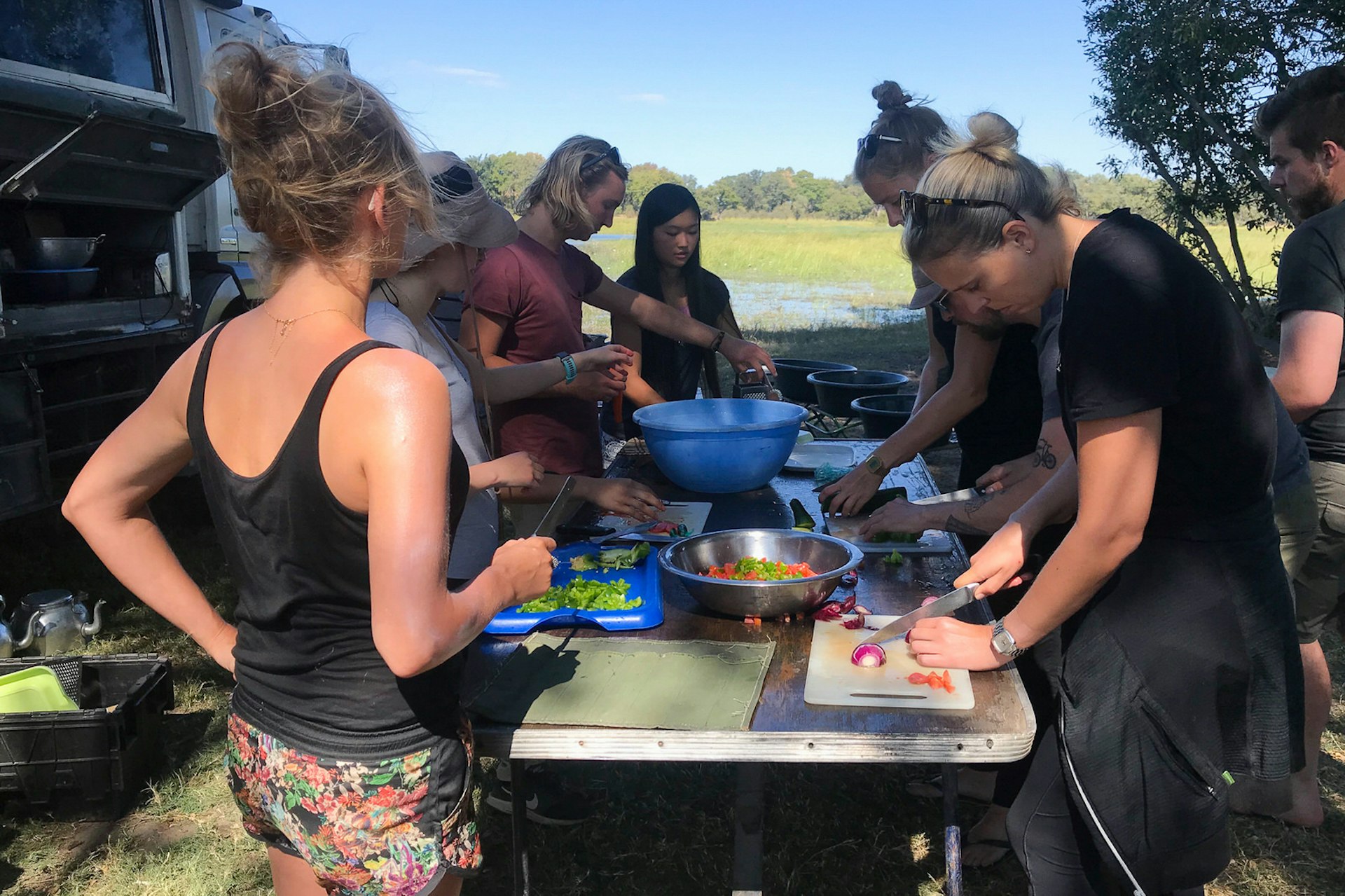 Half a dozen people are standing around a portable table, each preparing something different for an upcoming meal; the overland truck sits next to them in the wilderness @ Sarah Reid / ϲʼʱ