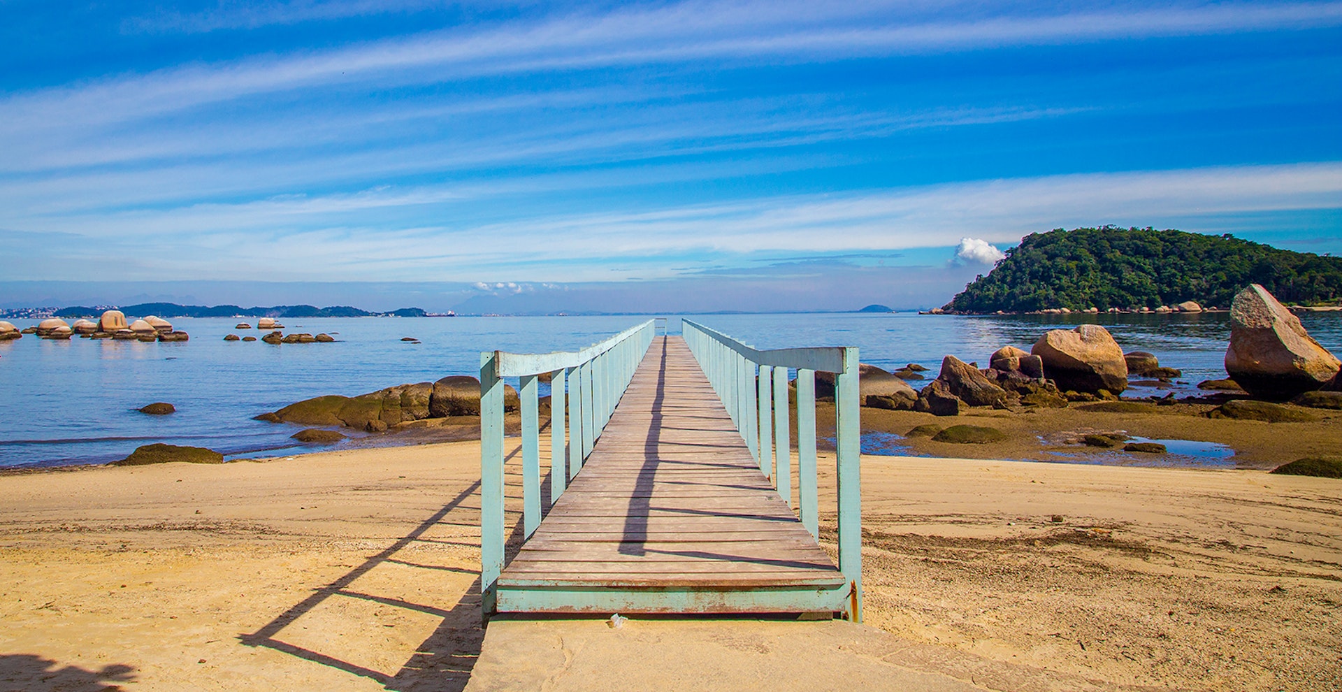 A wooden pier painted teal stretches off a rocky coastal beach in Guanabara Bay, Ilha da Paquetá, Brazil.
