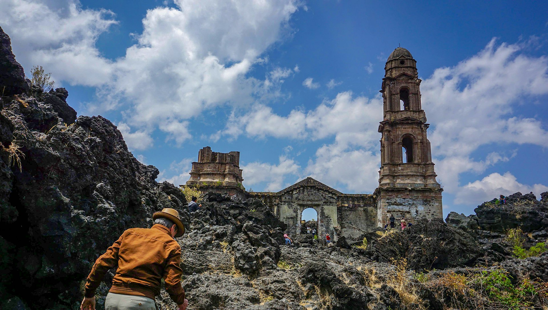 A man in an orange jacket and fedora walks towards a church ruin in a lava field in Central Mexico