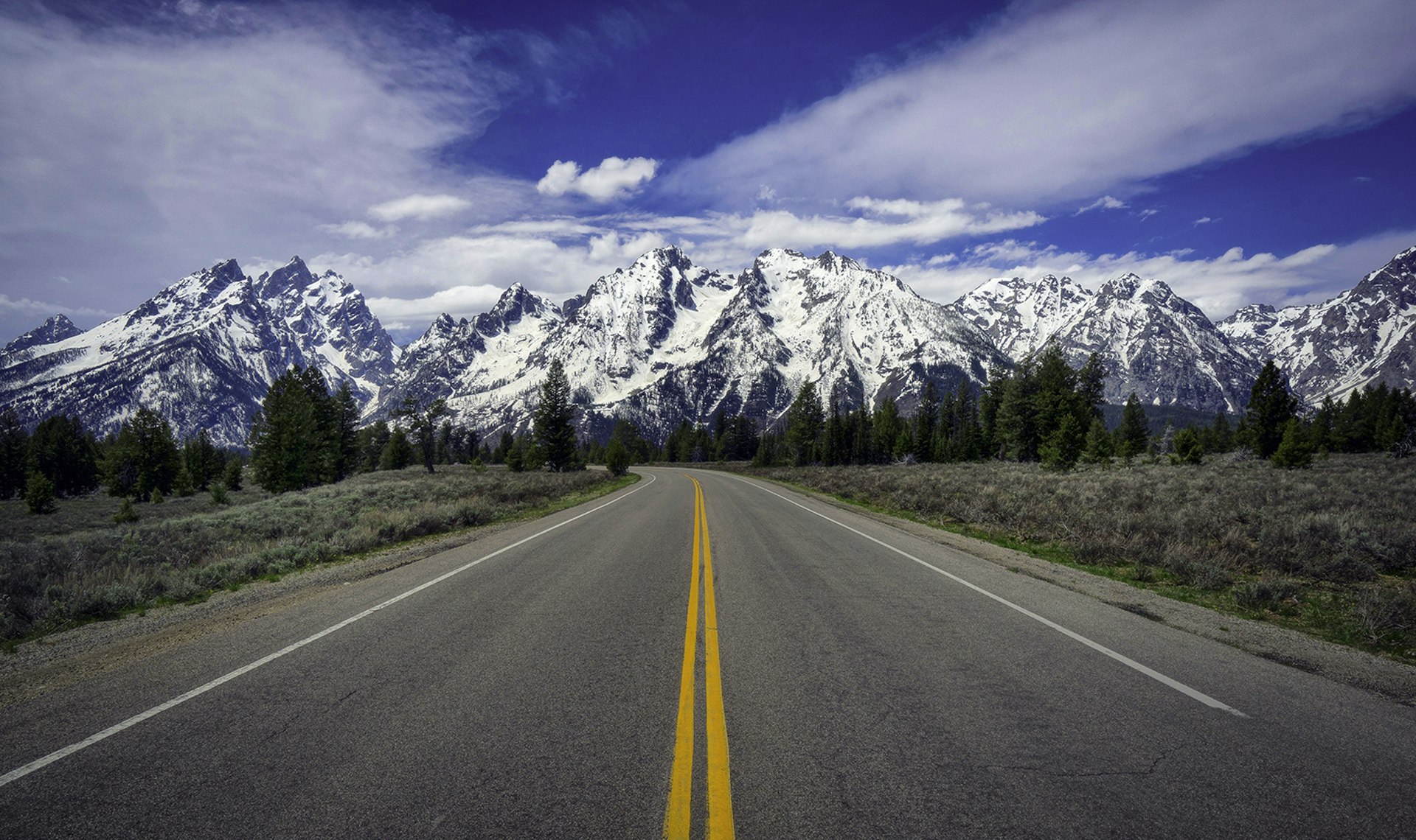 A single-lane road stretches straight forward towards snowy mountain peaks in the Rocky Mountains, USA.