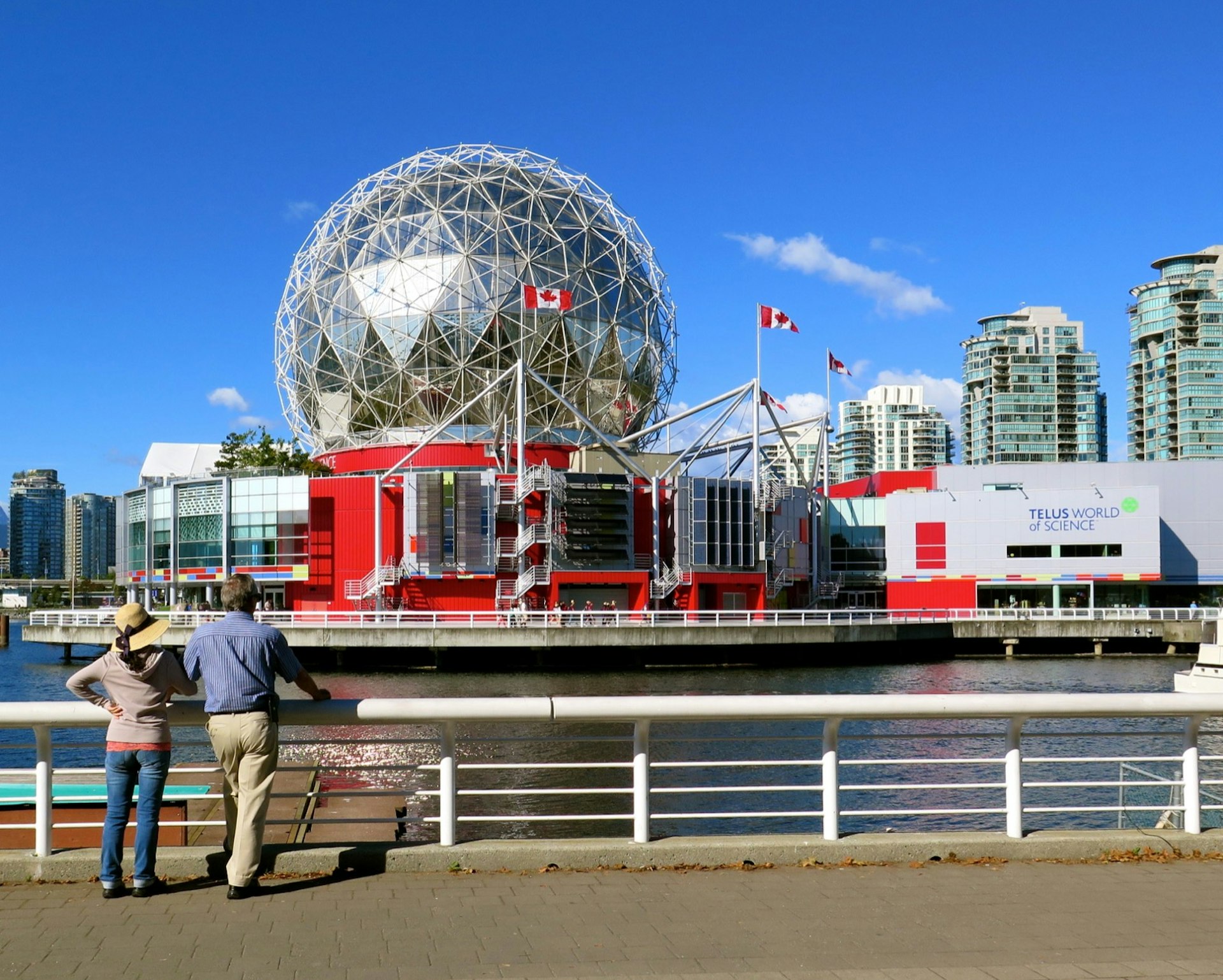 the glass dome of the Science World museum, with fluttering Canada flags, is seen across a waterway as tourists look at it from a dock