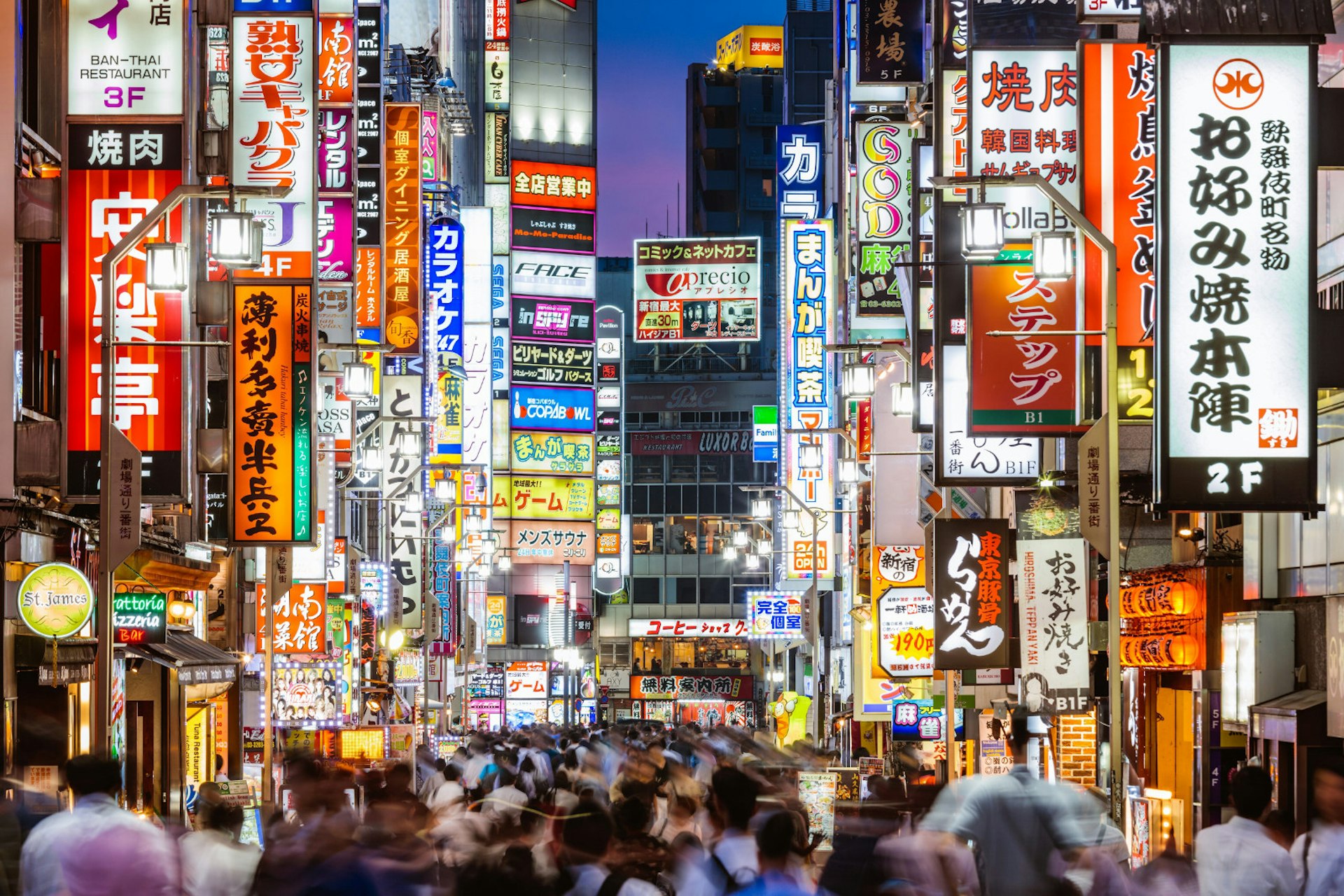 The bustling Tokyo neighbourhood of Shinjuku at night; a huge crowd of people walk under a mass of neon-lit signs in Japanese script.