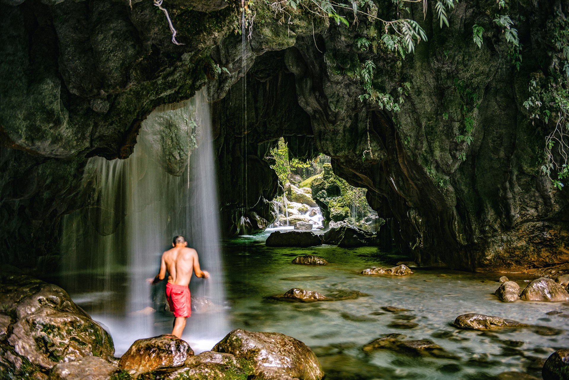 A man in red shorts stands under a waterfall within a cave tunnel in the Sierra Gorda, Mexico