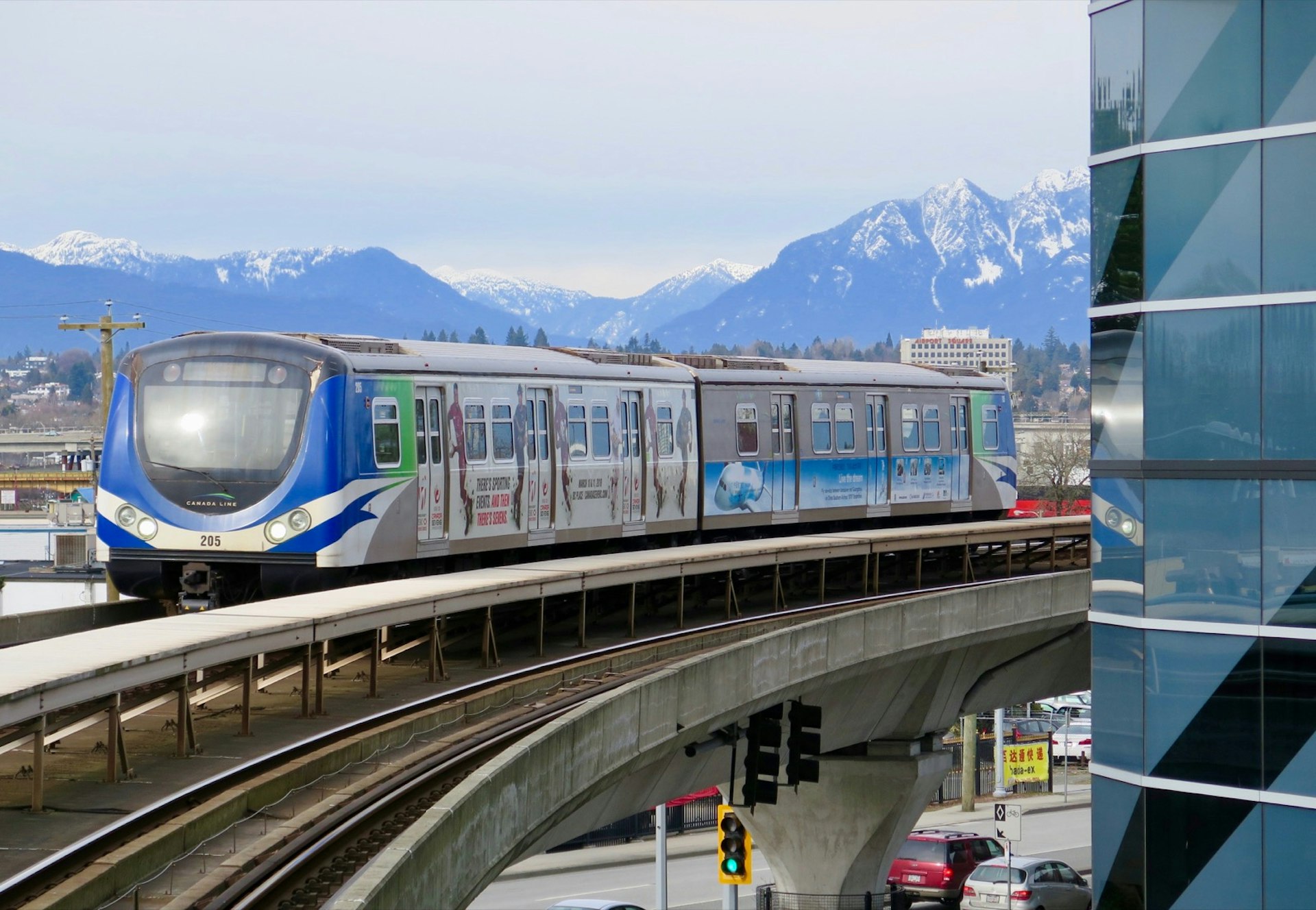 A blue and white and silver SkyTrain train on an elevated track curves around a building with a range of snow-covered mountains in the background