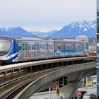 A blue and white and silver train on an elevated track curves around a building with a range of snow-covered mountains in the background