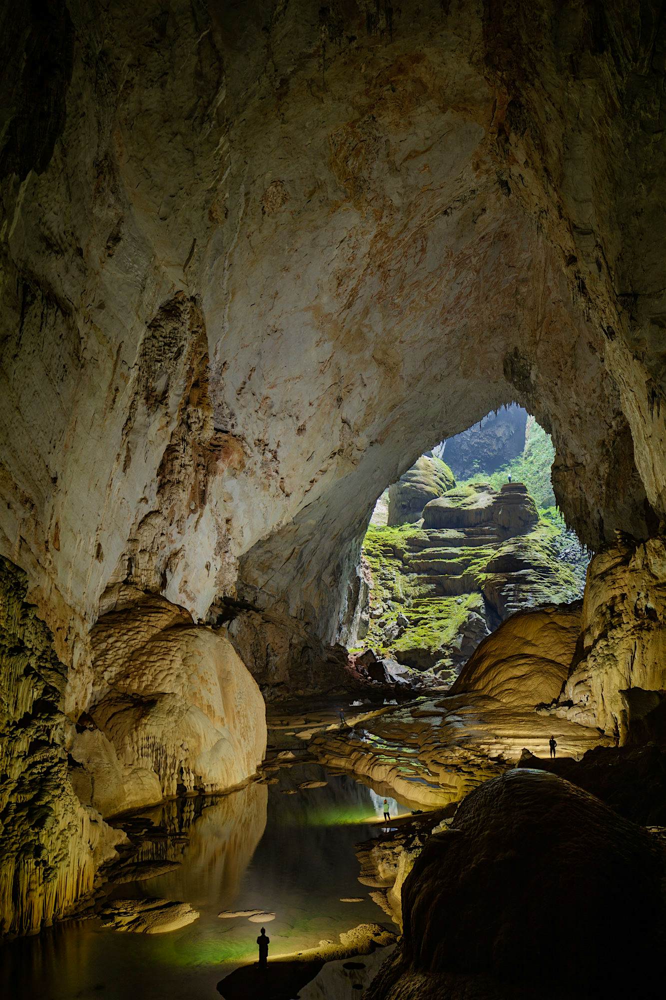 A large cave with water and boulders near the entrance. The size of this cave is put into context by the silhouettes of several people standing at various points within the cave