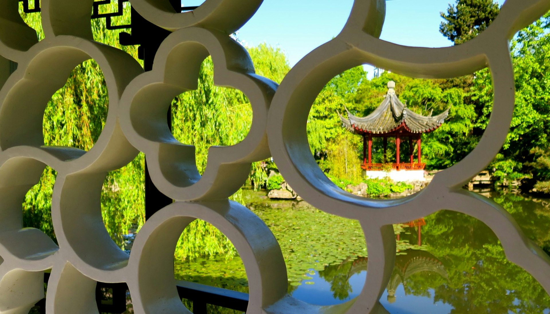 A red pagoda on a pond is seen through the ornate stone scrollwork on a bridge bannister