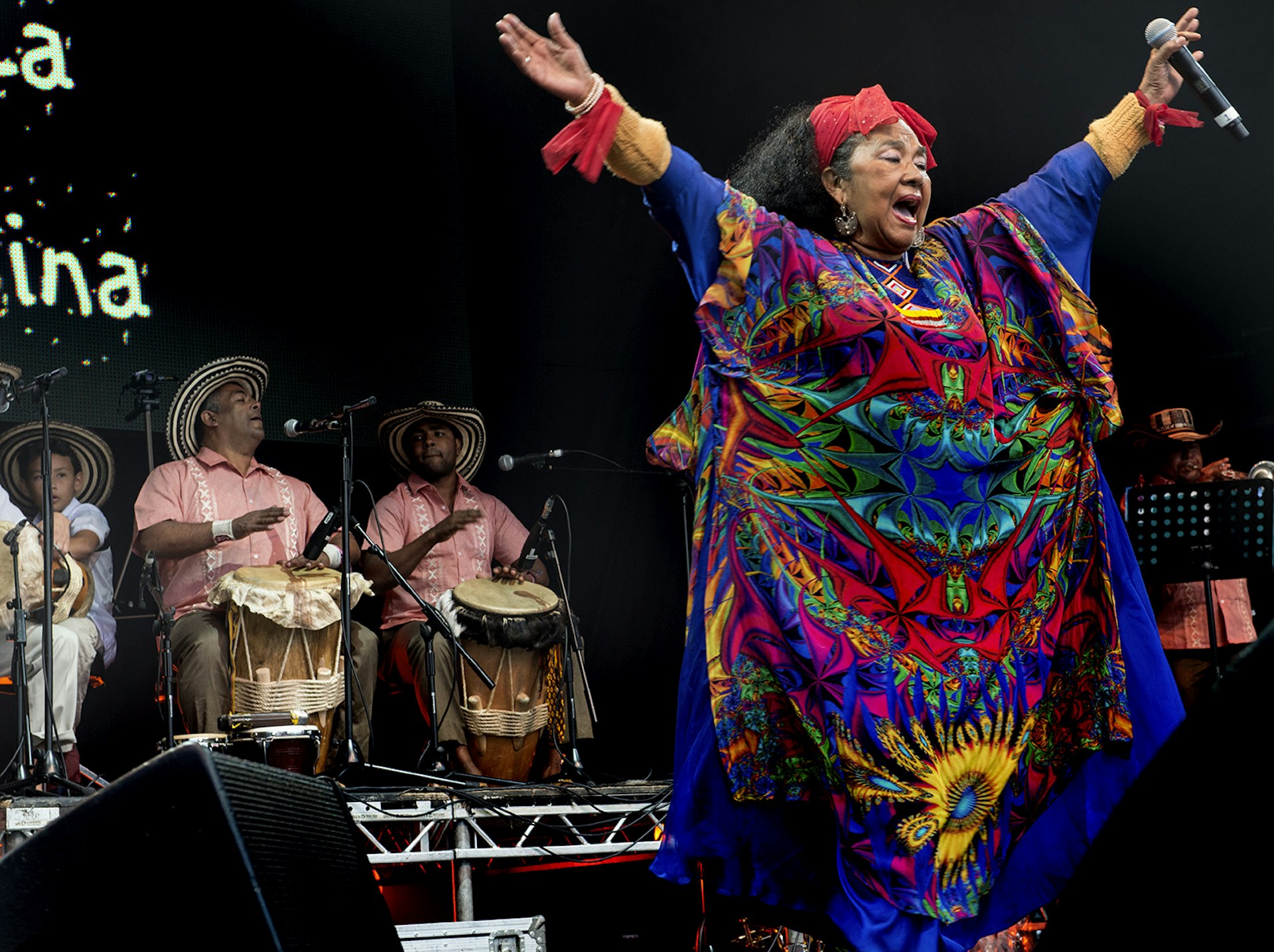 A singer wearing a colorful, flowing dress raises her hands to the crowd while men wearing wide-brimmed hats play the drums behind her