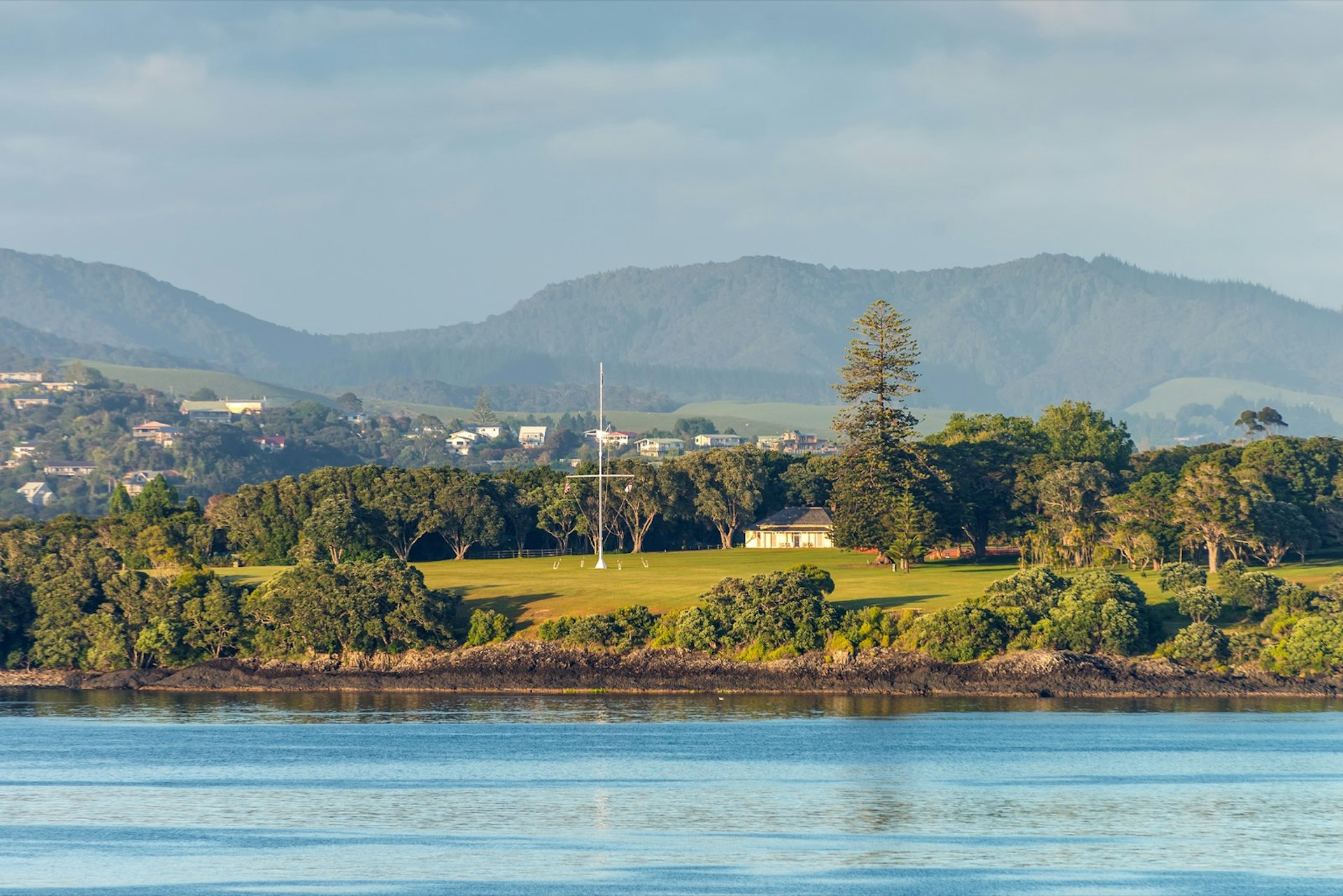 In the foreground are calm ocean waters; they abut a rocky, tree-lined shore that makes way to a lush lawn that is home to a large flagpole and single building nestled among trees; forested hills dotted with homes fill the background; the scene is the Waitangi treaty grounds in Paihia, Northland, New Zealand