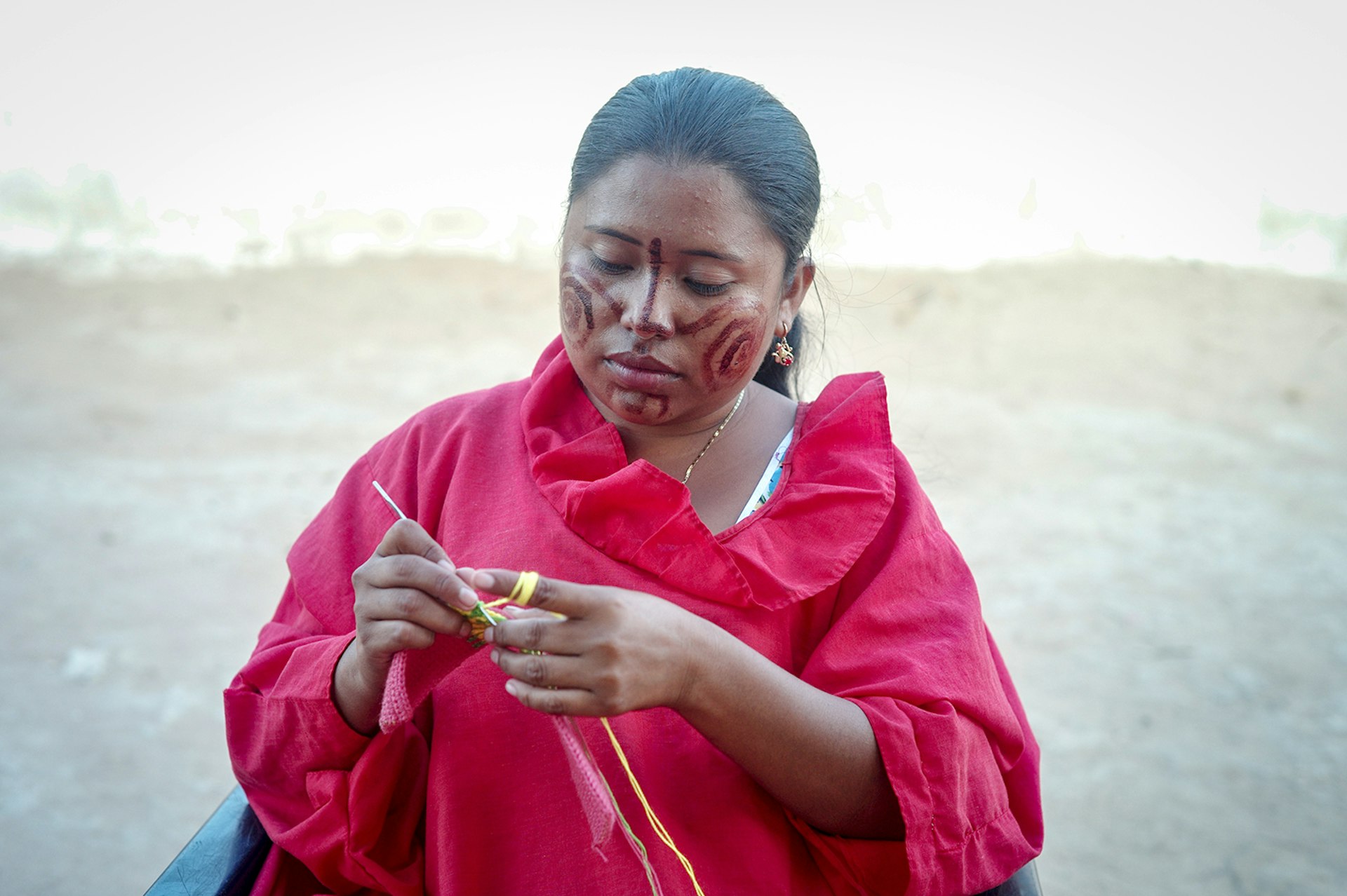 A Wayúu woman wearing a red robe and red face paint weaves a small piece of fabric 