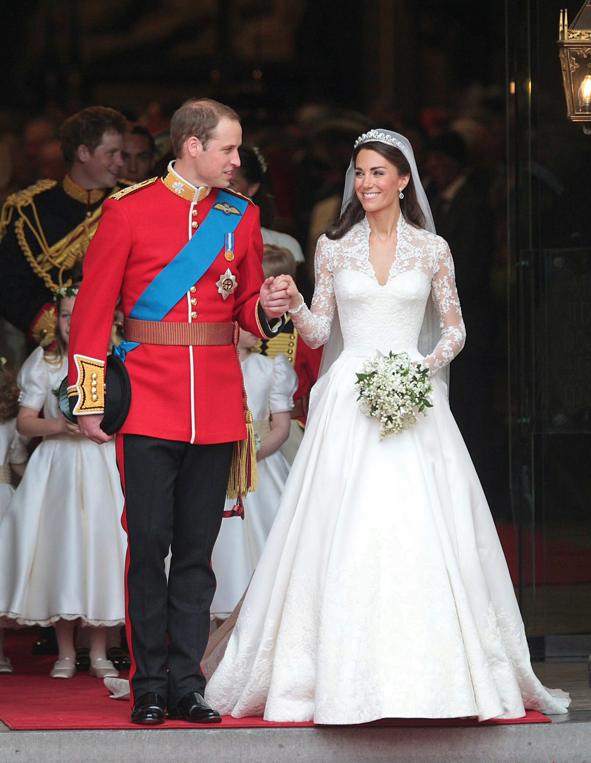 Prince William adorned in his ceremonial military uniform (bright red jacket, with embroidered gold lapels and blue sash, atop black trousers) smiles and holds the hand of Catherine who is wearing a beautiful white wedding dress with lace shoulders; a royal wedding to remember © Lewis Whyld / PA Images / Getty Images