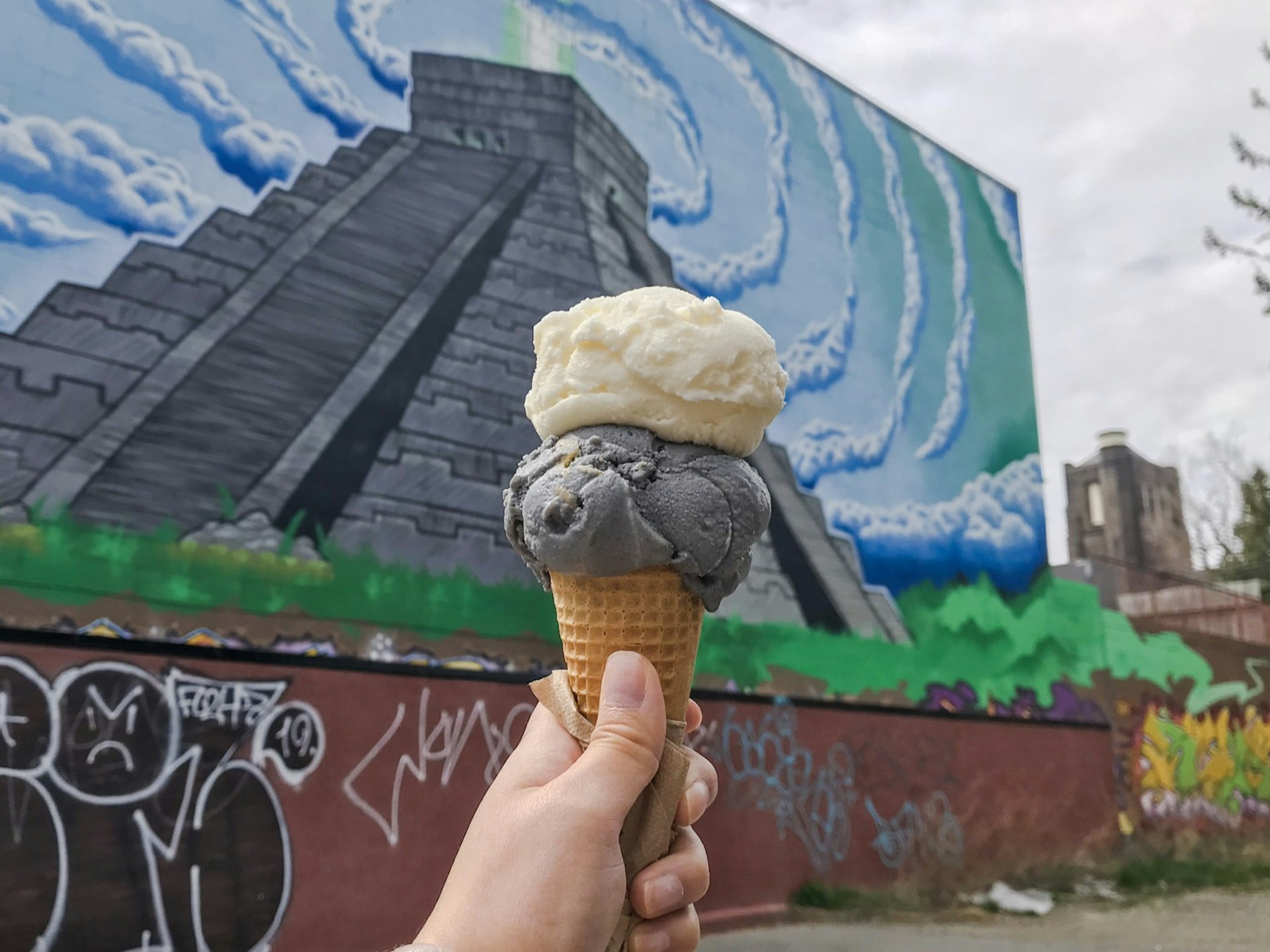 A hand holds two scoops of ice cream on a cone in front of a mural depicting a Mexican pyramid