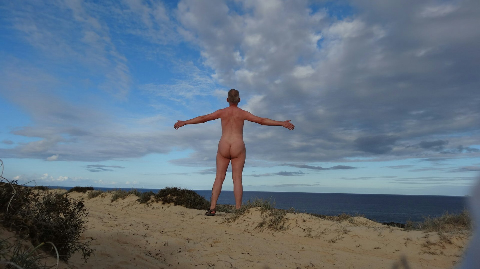With arms raised by his side, a nude man stands atop a sandy dune on a nude beach and faces the not-too-distant sea; the blue sky above has a beautiful swirl of white and grey clouds