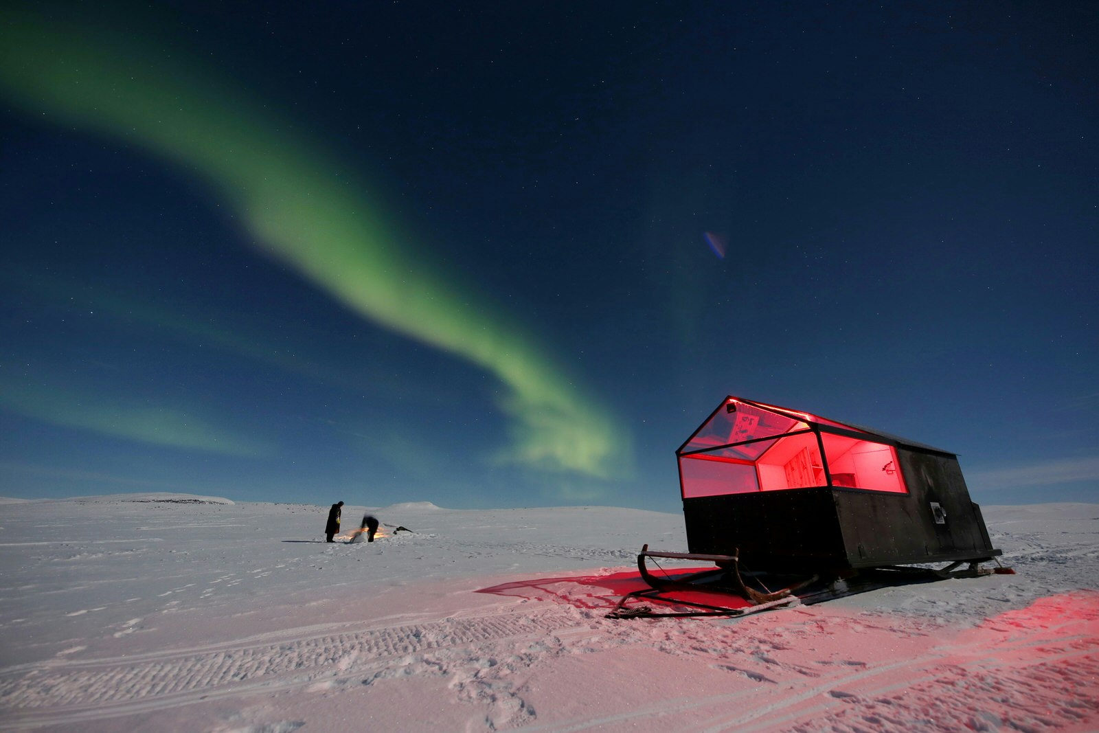 A small hut with a clear roof stands on large skis in the middle of a snowy, bare landscape with the Northern Lights in the sky and two people standing the background.