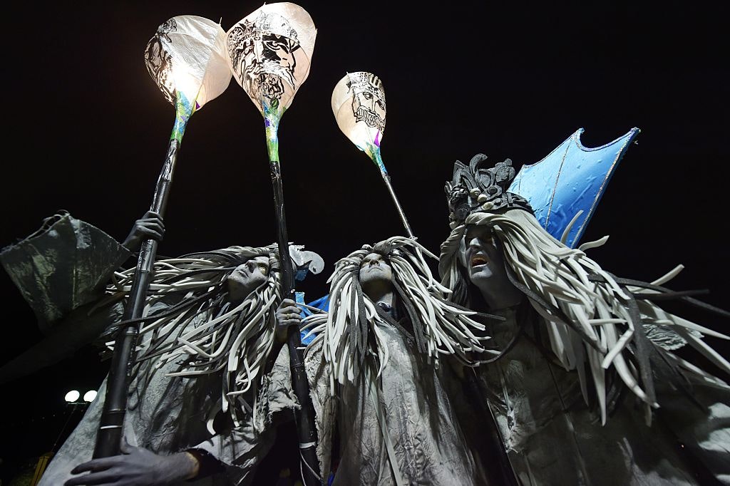 Three performers with grey robes and hair and blue wings hold lanterns during the night time parade.