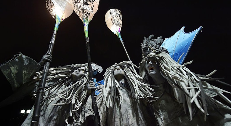 Three performers with grey robes and hair and blue wings hold lanterns during the night time parade.