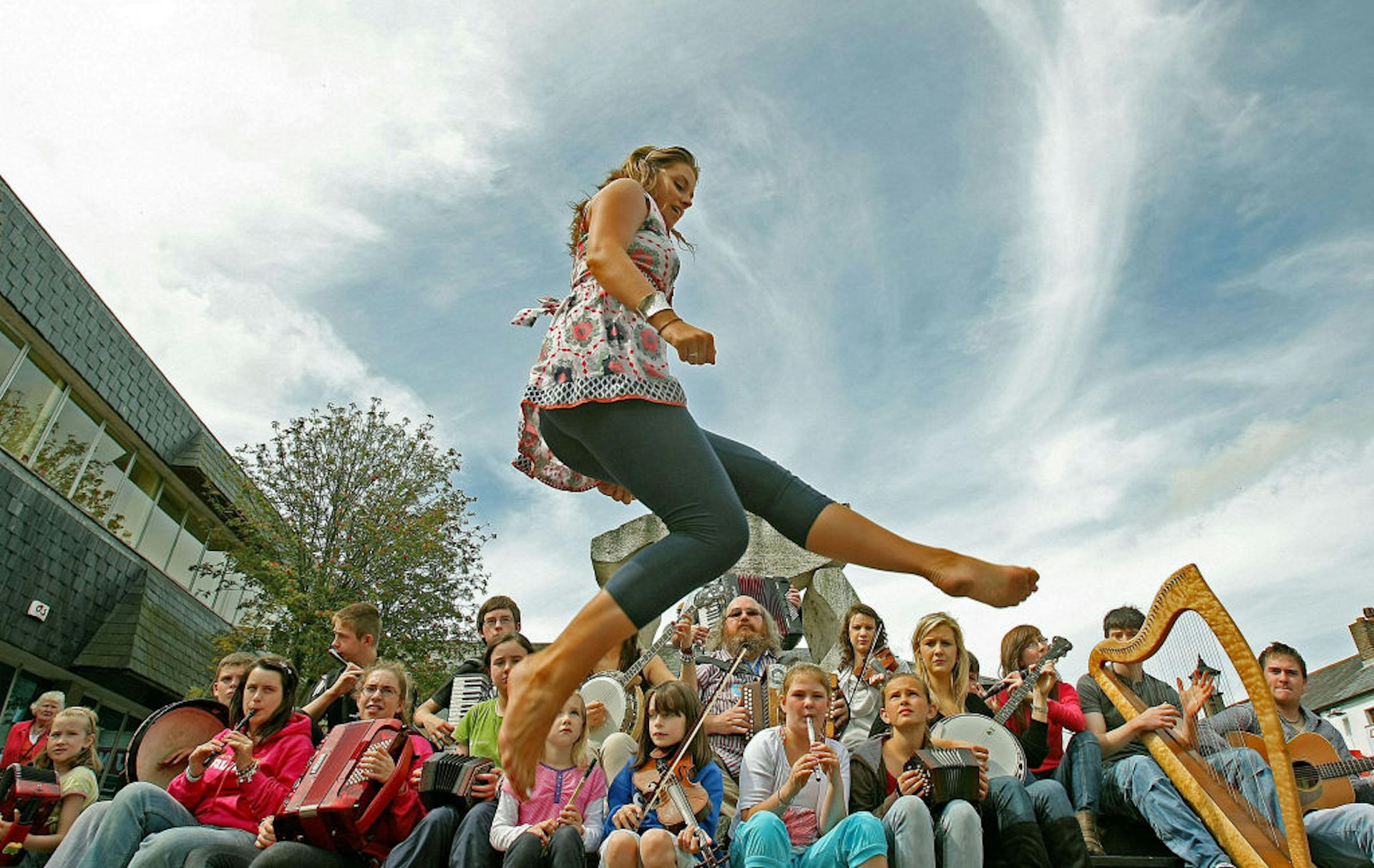 An Irish dancer in mid leap on a sunny day, with a crowd of young musicians playing traditional Irish instruments in the background.