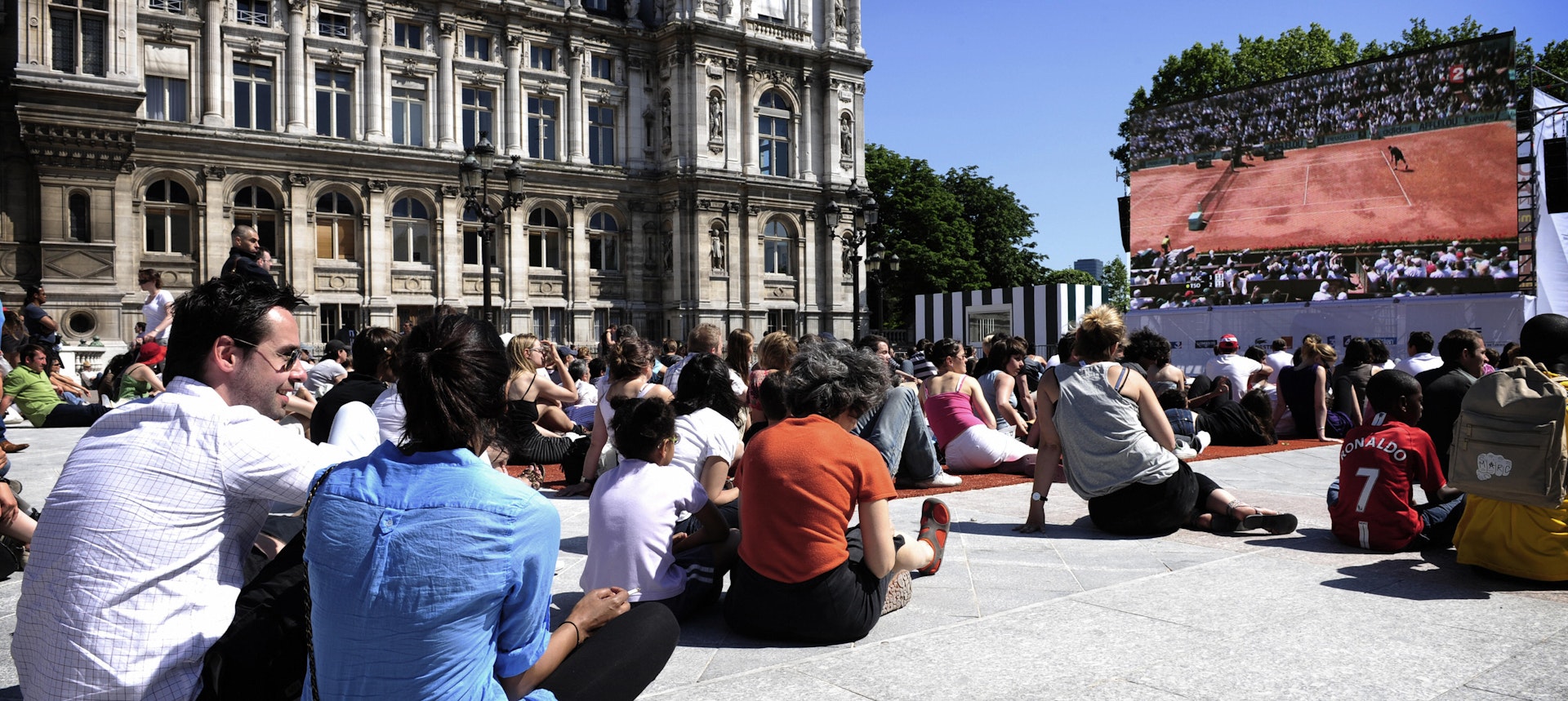 A large crowd sits in the sun next to the beautifully-ornate facade of the city hall while watching the French Open tennis on a huge screen