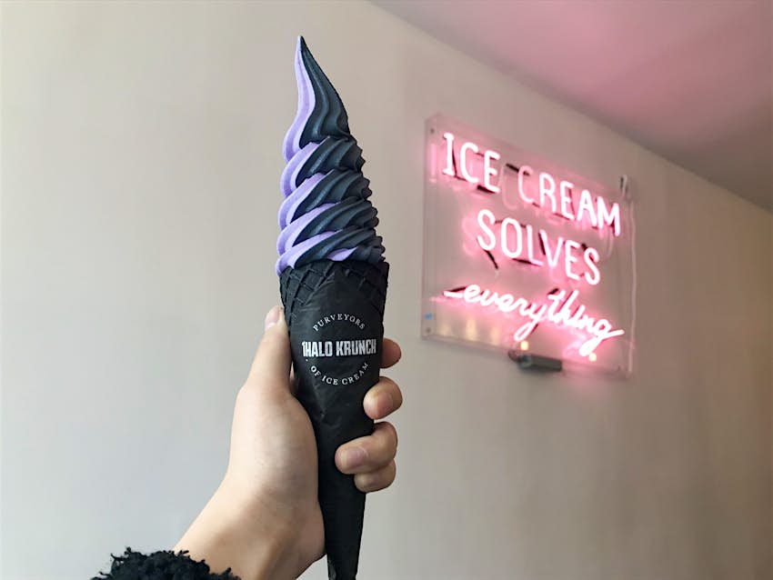 An intimidating-looking ice cream cone - black swirls of soft serve in a black cone - is held in front of a pink neon sign that reads ice cream solves everything