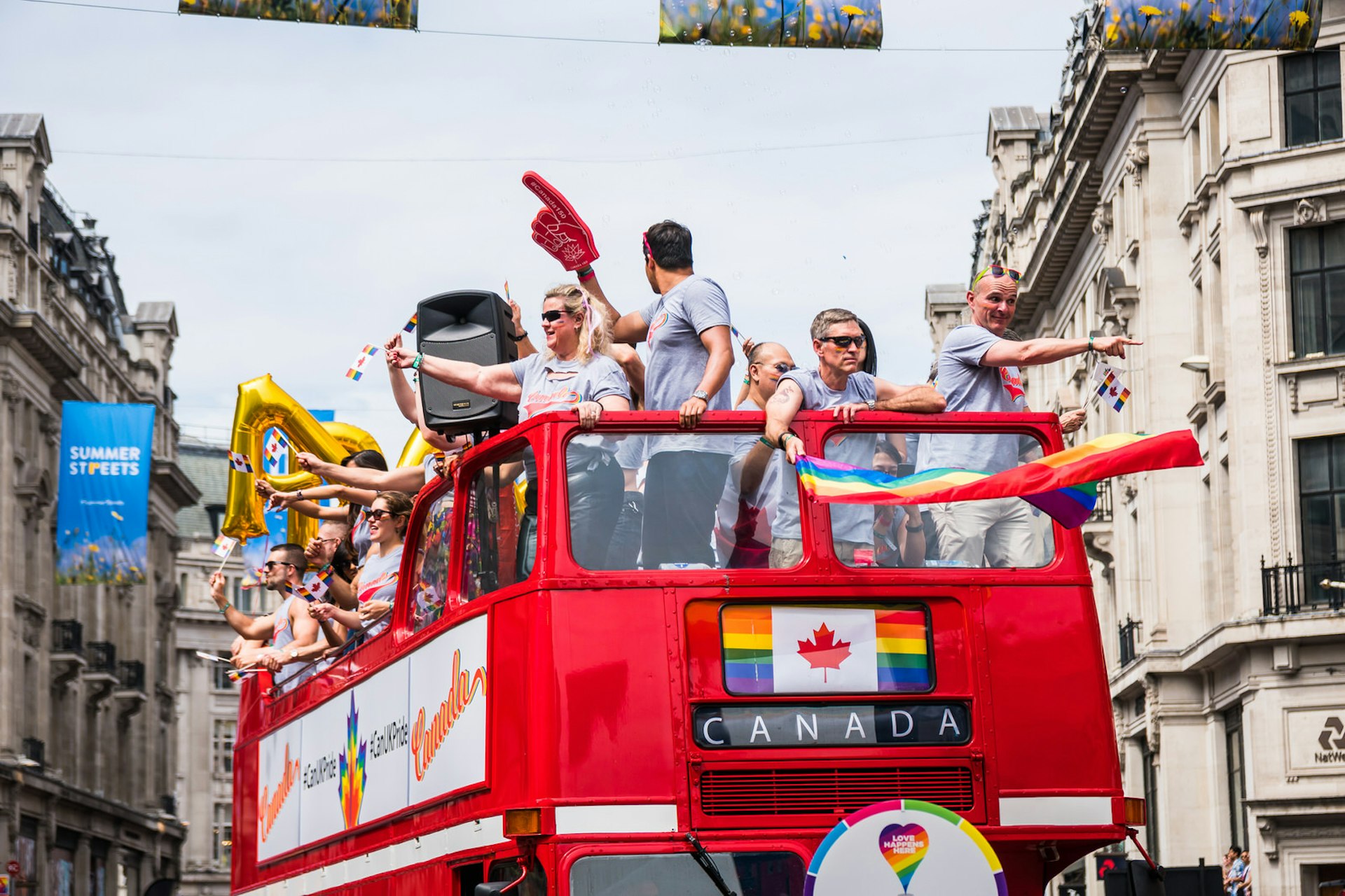 People on a topless double decker red London bus on the Pride in London parade on Regent Street; the bus' destination reads CANADA and there is a Canadian flag above it, with the normally red stripes replaced by rainbow colours