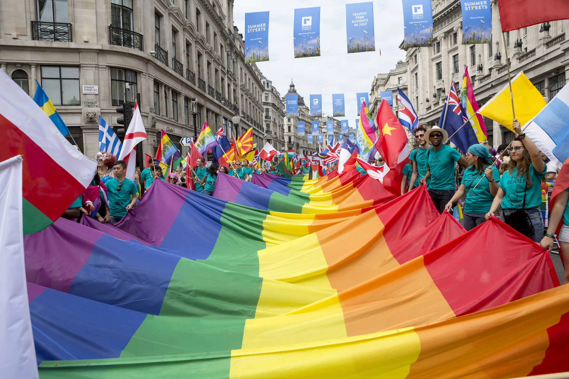 Pride in London parade participants carrying a huge rainbow flag along Regent Street; everyone is wearing matching green shirts that say 'flag bearer' on them, and unsurprisingly, they each have one hand on the rainbow flag and the other on their nation's flag (those of Sweden, Greece, China, New Zealand, Belarus are visible) 