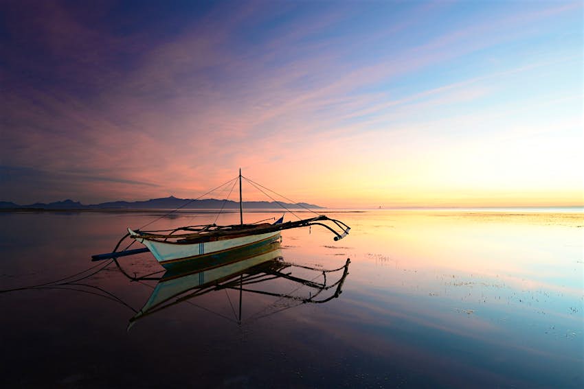 A traditional bangka (lightweight outrigger boat) sits on water so still it mirrors the pink-blue sunrise in the sky ©Julien Pons/500px