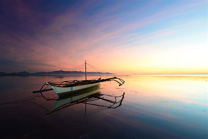 Features - palawan-travel-bangka A traditional bangka (lightweight outrigger boat) sits on water so still it mirrors the pink-blue sunrise in the sky ©Julien Pons/500px