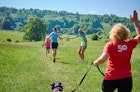 A child and youth run through green grasslands towards a forested hill; a supporter holds her hand out to the youth to slap as he passes, as a runner in the foreground heads away from the camera with her dog on a lead